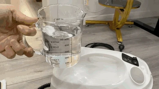 A hand holds a glass measuring cup filled with 500 milliliters of water positioned over the water filling opening of a white Kärcher SC3 steam cleaner.