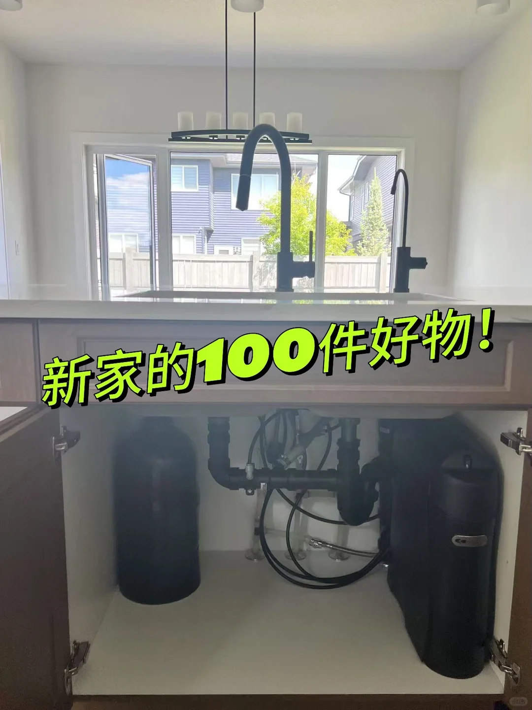 A kitchen island sink with an open cabinet underneath revealing black plumbing pipes and an under-sink water filtration system. Above the counter are two black gooseneck faucets set in front of a bright window.