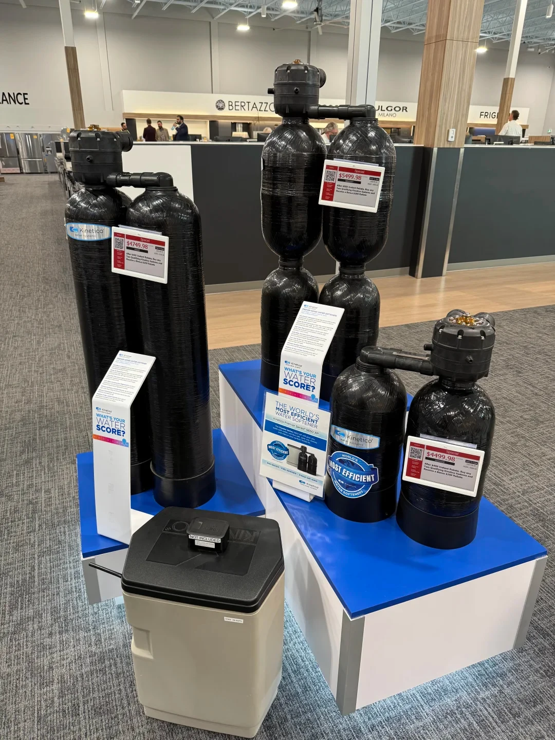 Display of three Kinetico water softener systems with black dual tanks in different sizes, accompanied by promotional placards, pricing signs, and a brine tank in a retail store showroom.