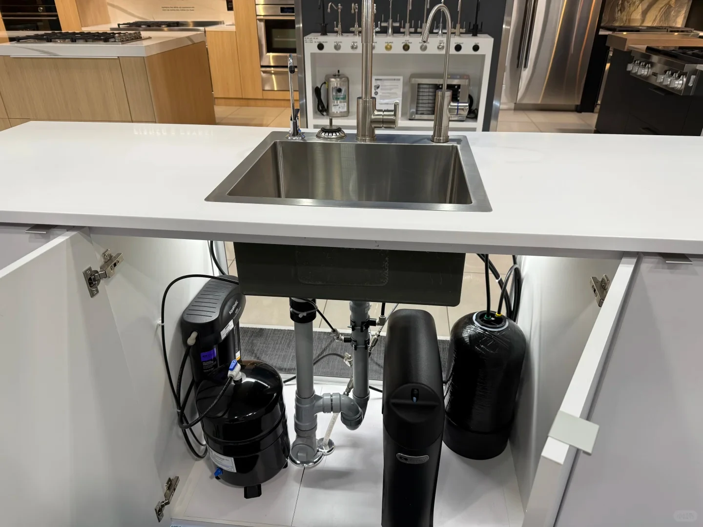 A kitchen island showroom display featuring a stainless steel undermount sink and faucet atop a white countertop, with the cabinet doors open below to reveal an installed under-sink water filtration system, storage tanks, and drainage plumbing.