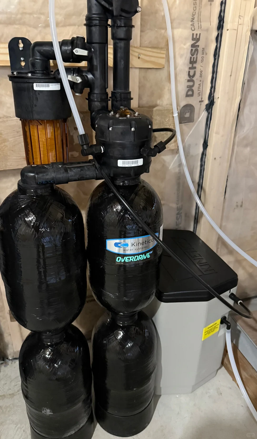 An installed Kinetico Overdrive water softening system featuring two tall black resin tanks, an amber pre-filter housing, connecting black and clear pipes, and a grey brine tank against an insulated basement wall.