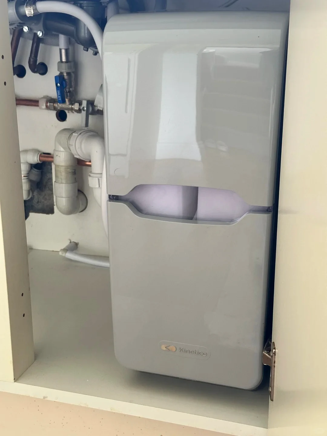 A gray Kinetico water softener unit installed inside an under-sink cabinet alongside plumbing pipes and valves.