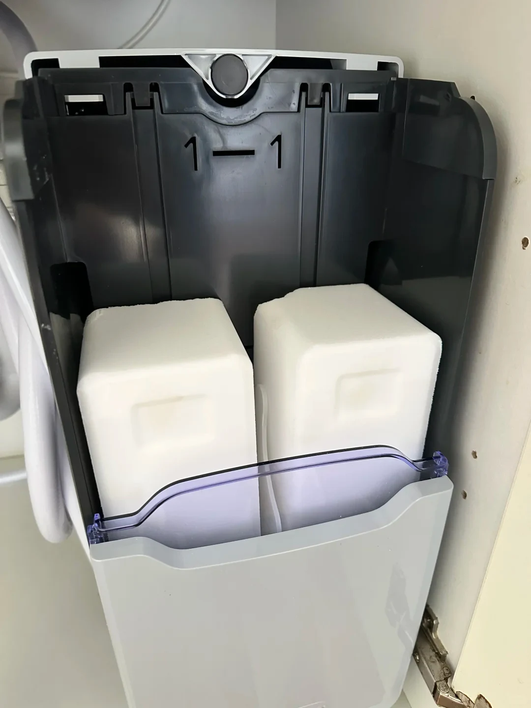 An open dark-grey salt compartment of a water softener installed inside a cabinet, holding two rectangular white salt blocks behind a light-grey front cover with a transparent purple rim.