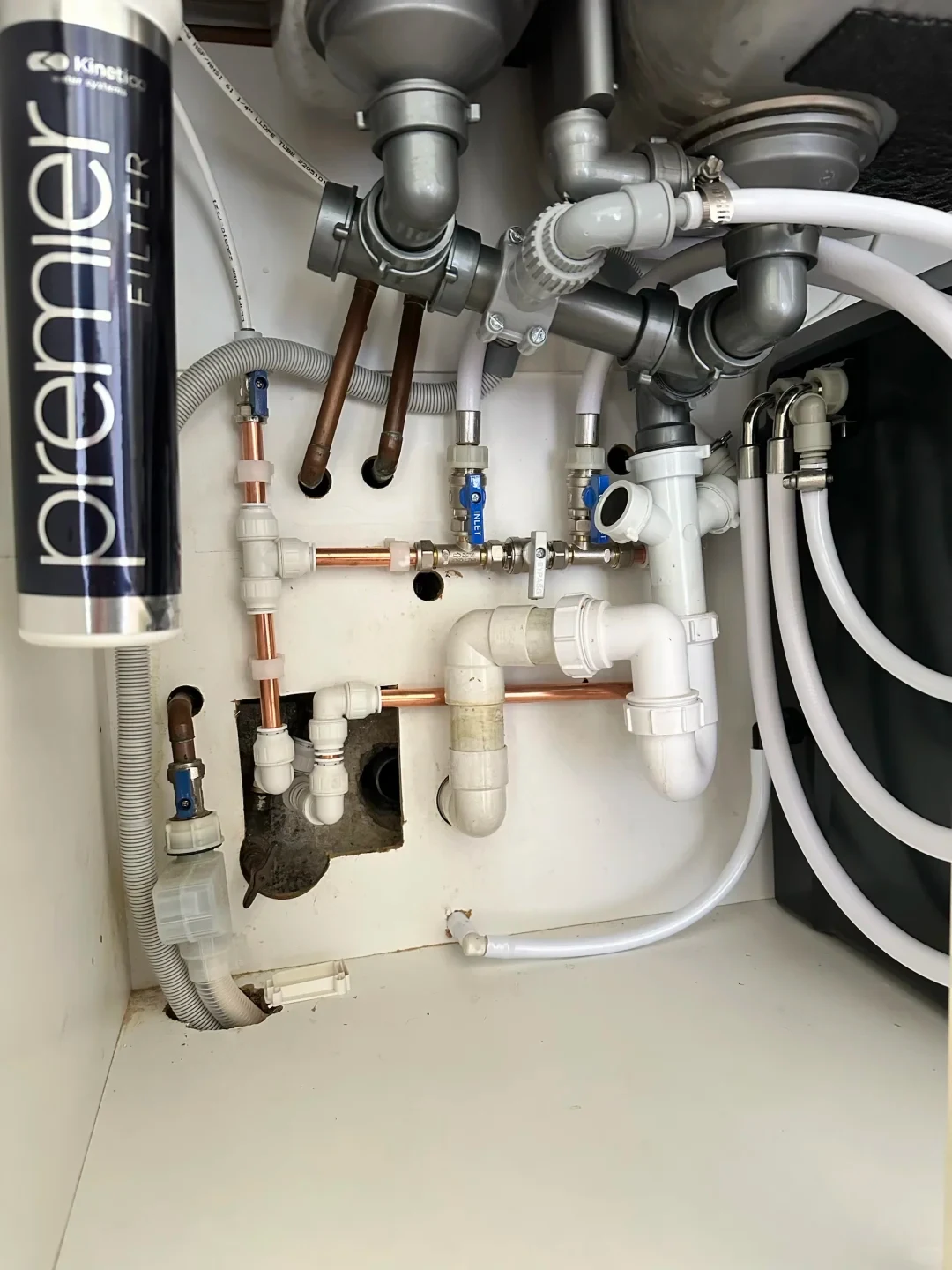 Under-sink plumbing setup with white and grey PVC drain pipes, copper water supply lines with blue lever valves, and a Kinetico Premier water filter cylinder installed on the left side.