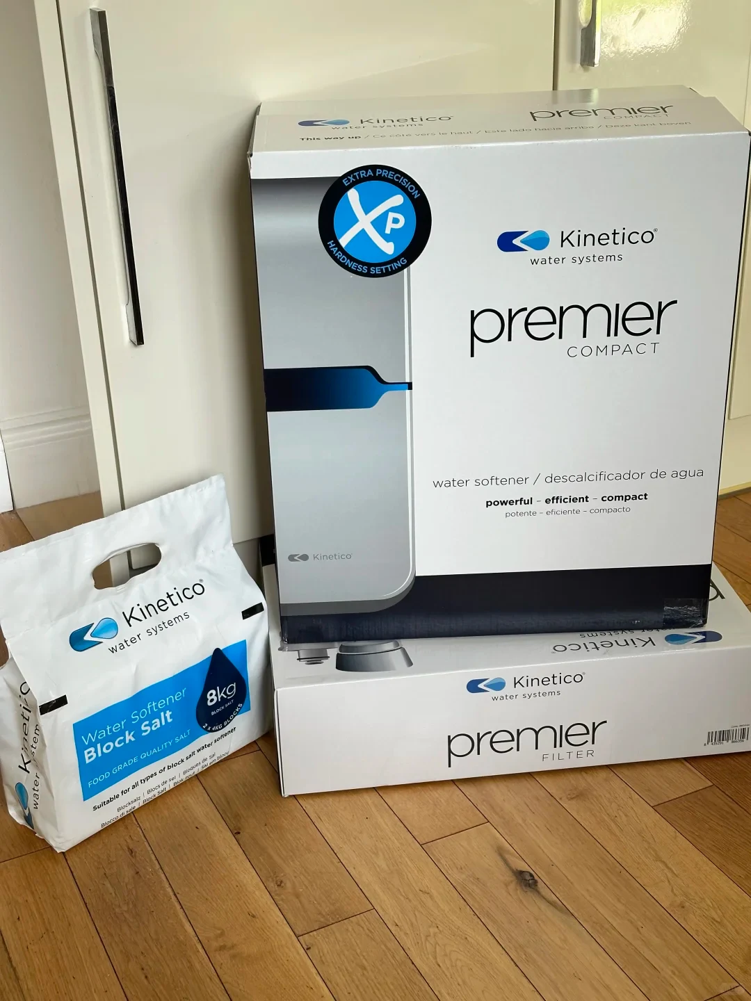 A Kinetico Premier Compact water softener box placed on top of a Kinetico Premier Filter box on a wooden floor, accompanied by an 8 kg bag of Kinetico Water Softener Block Salt.