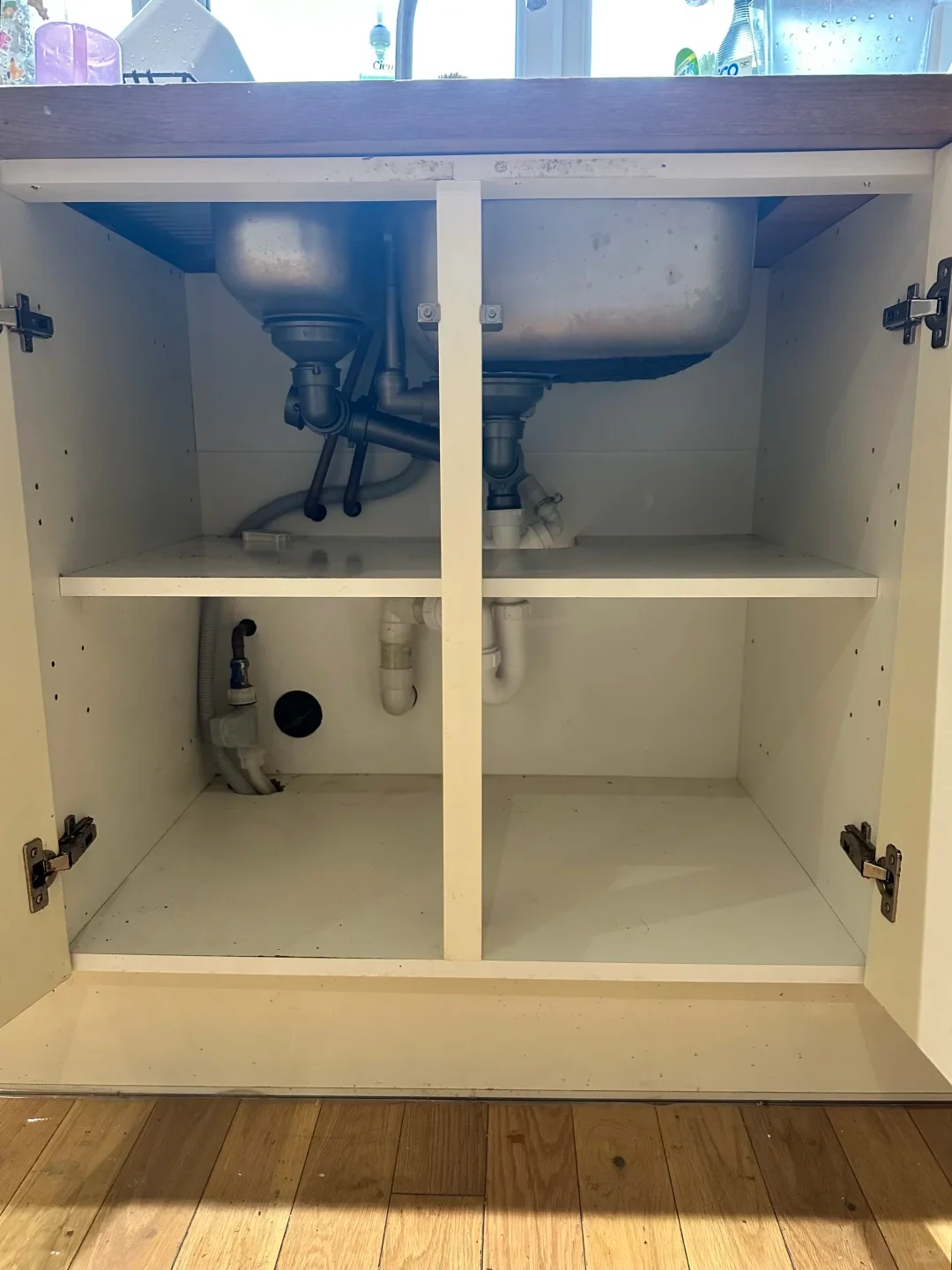 An open, empty white kitchen under-sink cabinet showing the stainless steel sink basin and attached plumbing pipes inside, divided horizontally by a white shelf with a central vertical post.