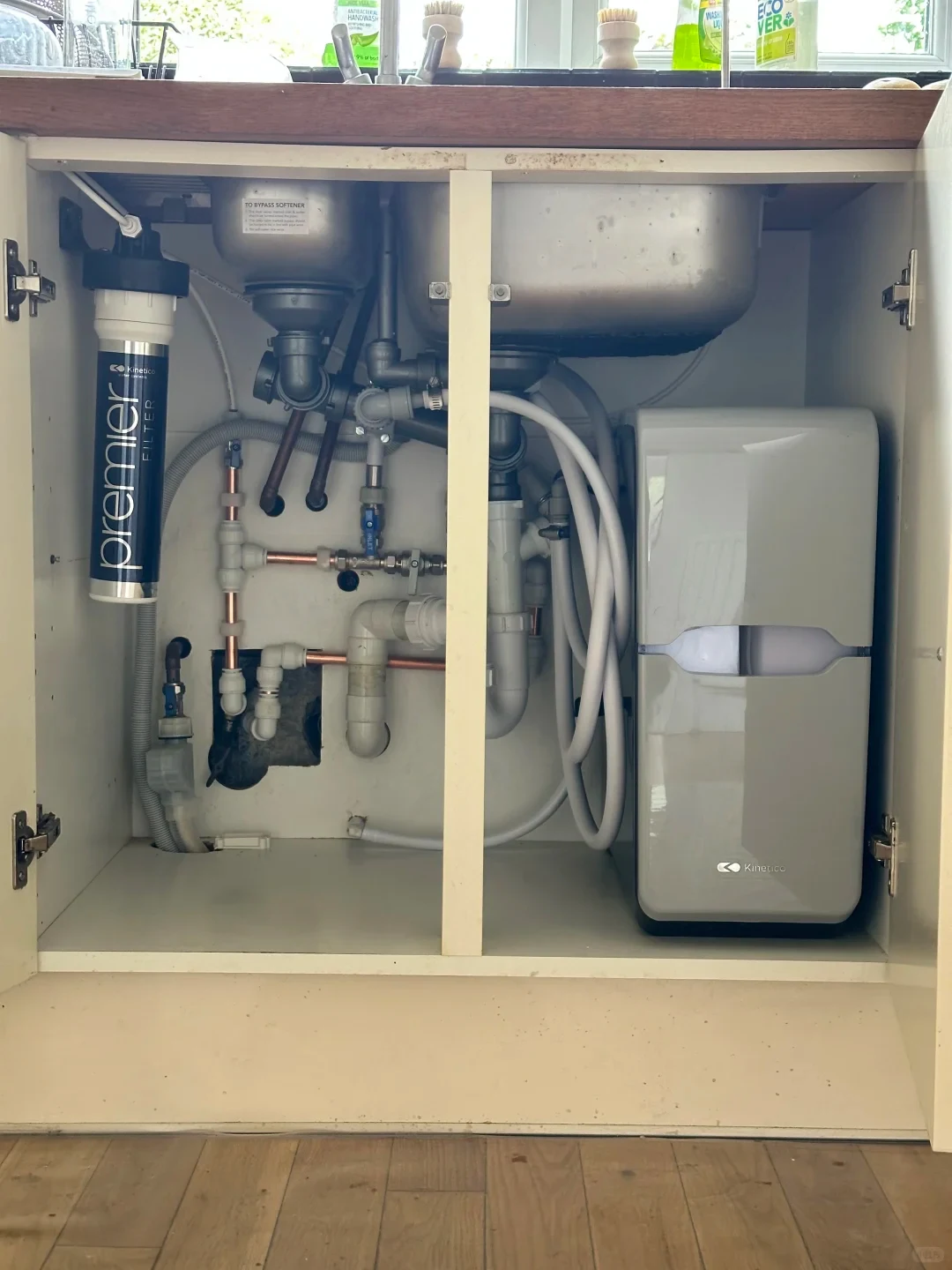 An open under-sink kitchen cabinet revealing an installed water treatment system and plumbing. Mounted on the left wall is a vertical black and white cylindrical Kinetico Premier water filter. On the right side sits a compact, grey Kinetico water softener unit with a semi-translucent viewing window. Various copper and PVC pipes, drainage traps, valves, and white flexible hoses connect the components beneath the stainless steel sink basin.