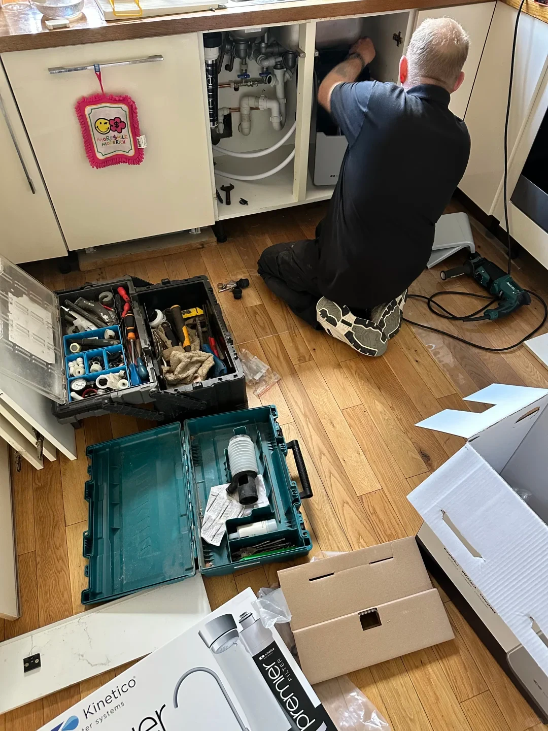 A technician kneels on a wooden kitchen floor while installing or servicing equipment inside an open base cabinet under the sink. Around him lie open toolboxes containing various tools and plumbing fittings, cardboard packaging, a Kinetico water filter system box, and a power drill.
