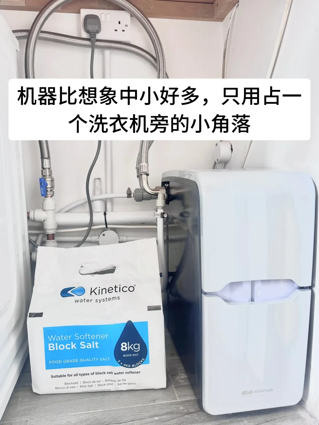 A compact white and grey Kinetico water softener installed in a tight space next to water pipes, beside an 8kg bag of Kinetico Water Softener Block Salt, with a caption banner overlaid at the top.