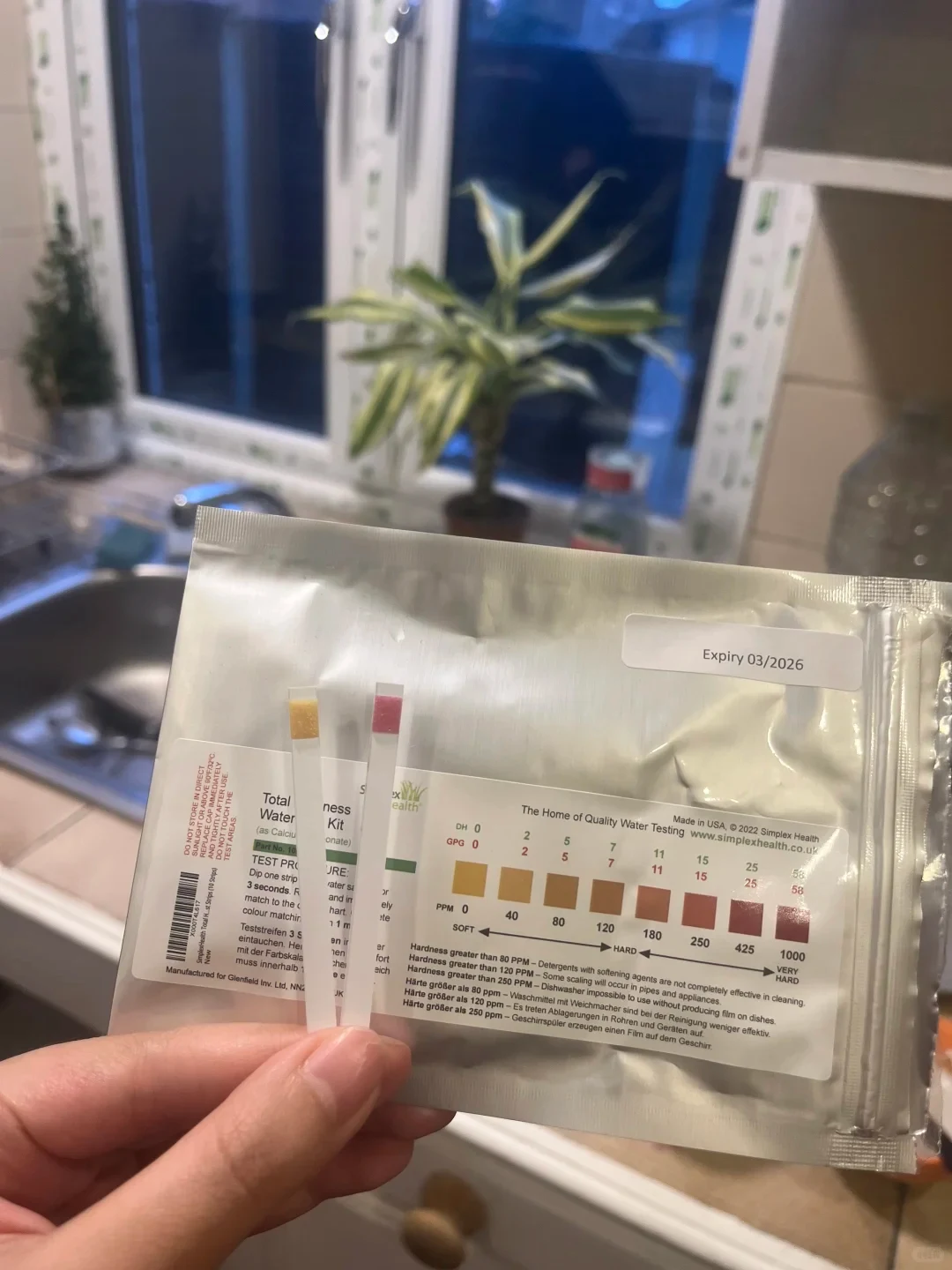 A hand holds two water hardness test strips in front of a SimplexHealth water testing pouch, comparing the strip colors to a soft-to-hard PPM chart above a kitchen sink.