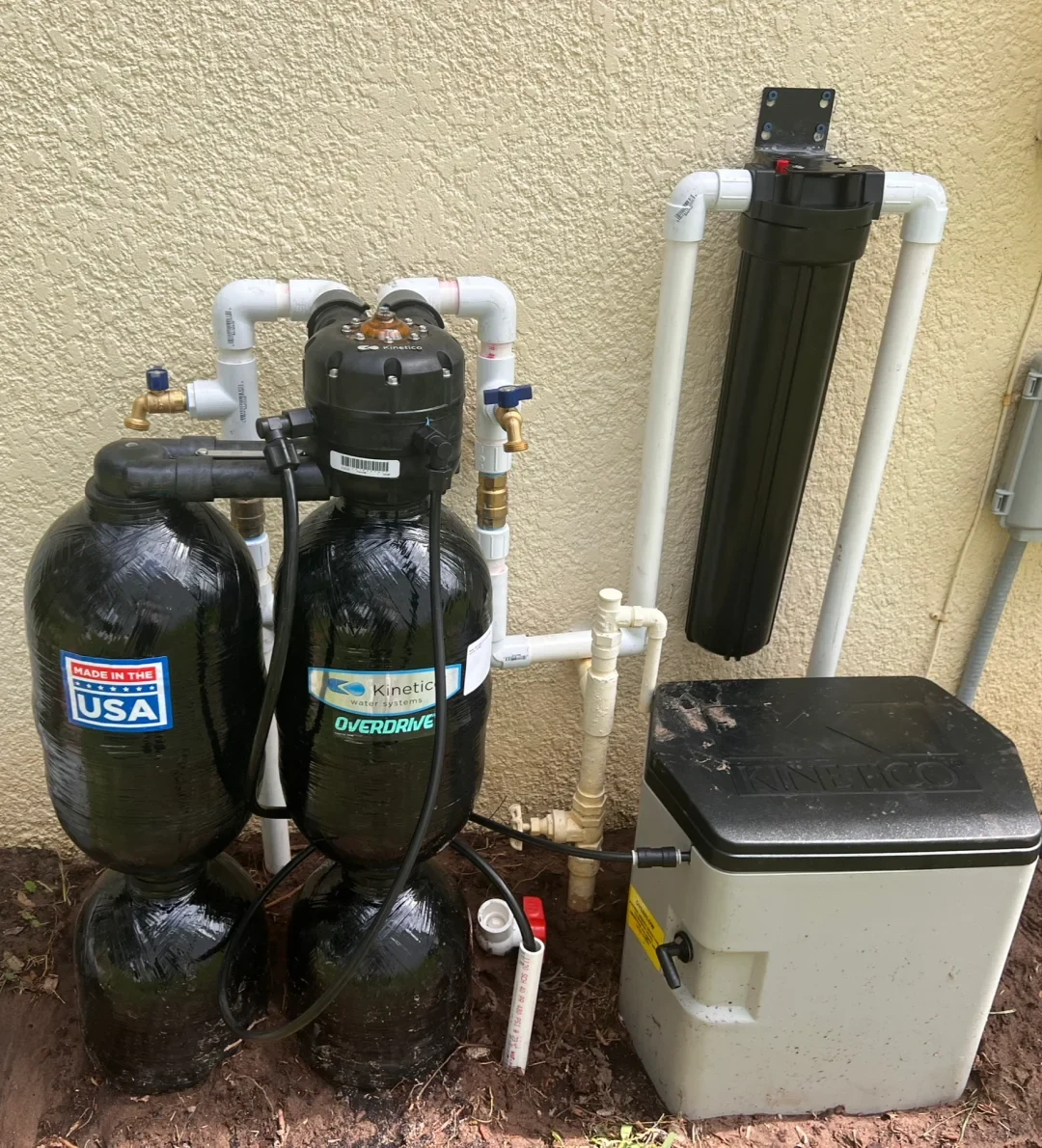 Outdoor residential water softening and filtration system installed beside a stucco exterior wall, featuring dual black Kinetico tanks, an upright pre-filter canister, and a brine tank connected by white PVC pipes.