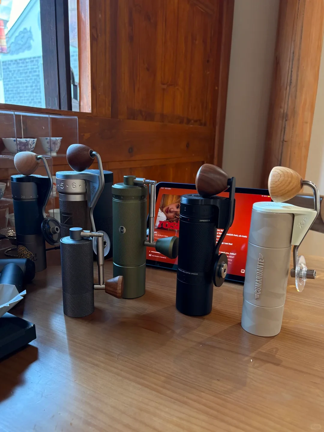 Several manual coffee grinders in different colors and sizes are displayed on a wooden table, with a tablet playing a video behind them.