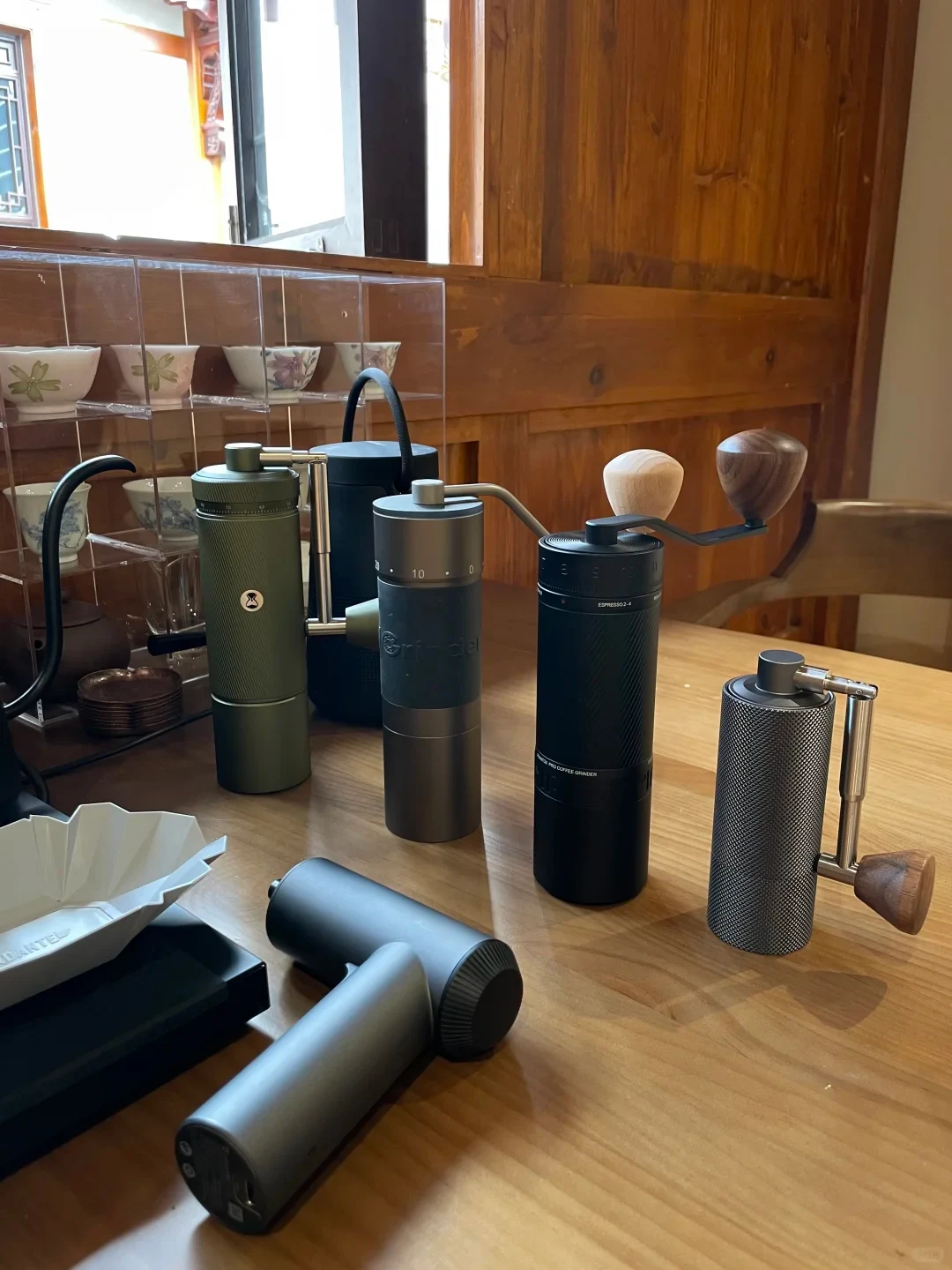 Several manual coffee grinders in different colors and sizes are displayed on a wooden table, with ceramic cups on shelves behind them.