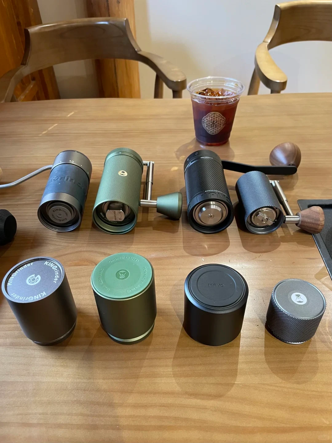 Several manual coffee grinders in different colors and sizes are arranged on a wooden table, with a clear cup of iced coffee behind them and wooden chairs in the background.