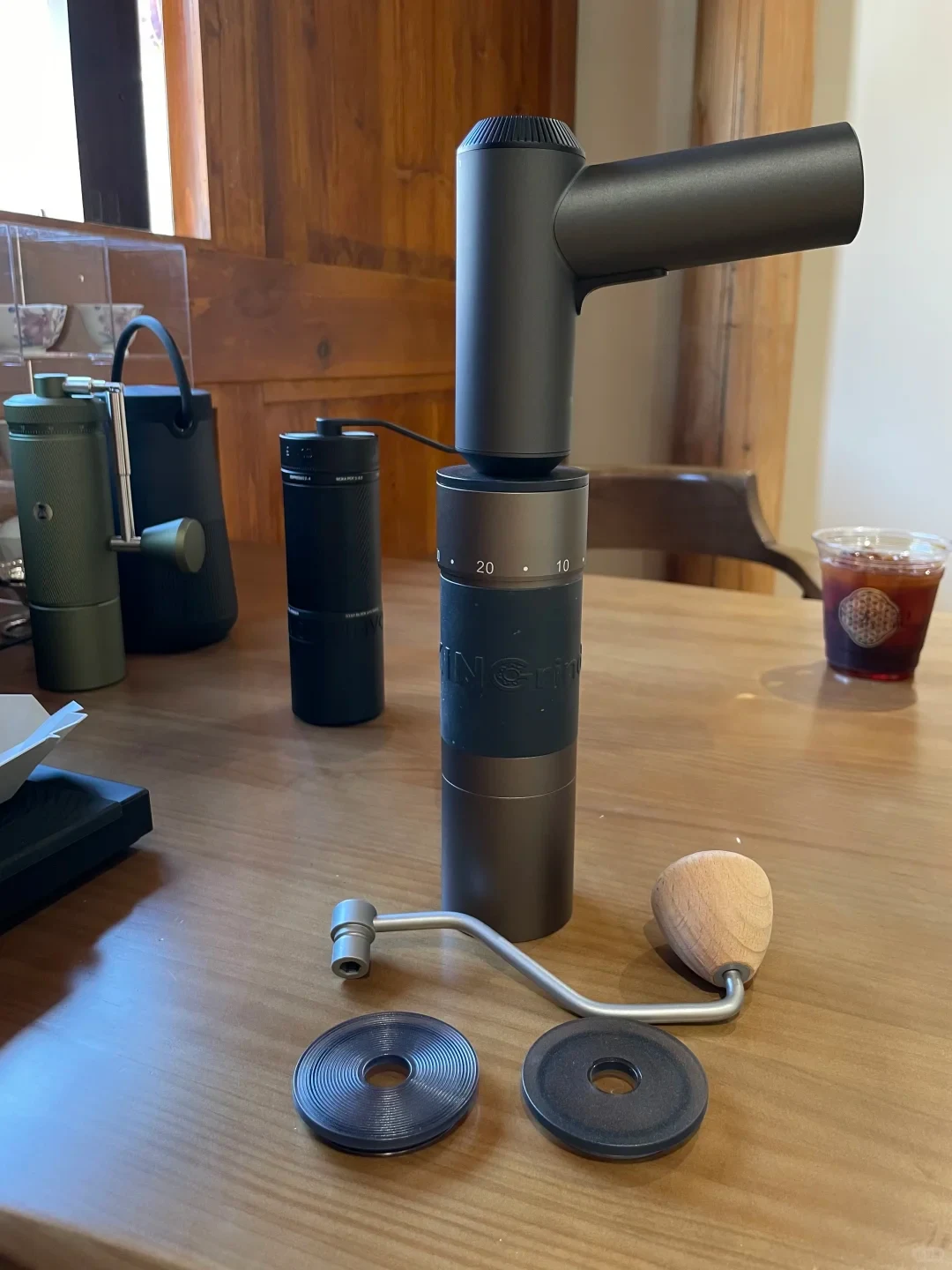A compact electric driver is mounted on top of a KINGrinder K6 coffee grinder on a wooden table. The removed crank with a wooden knob and two round lid parts lie in front; other grinders and an iced drink sit behind it.