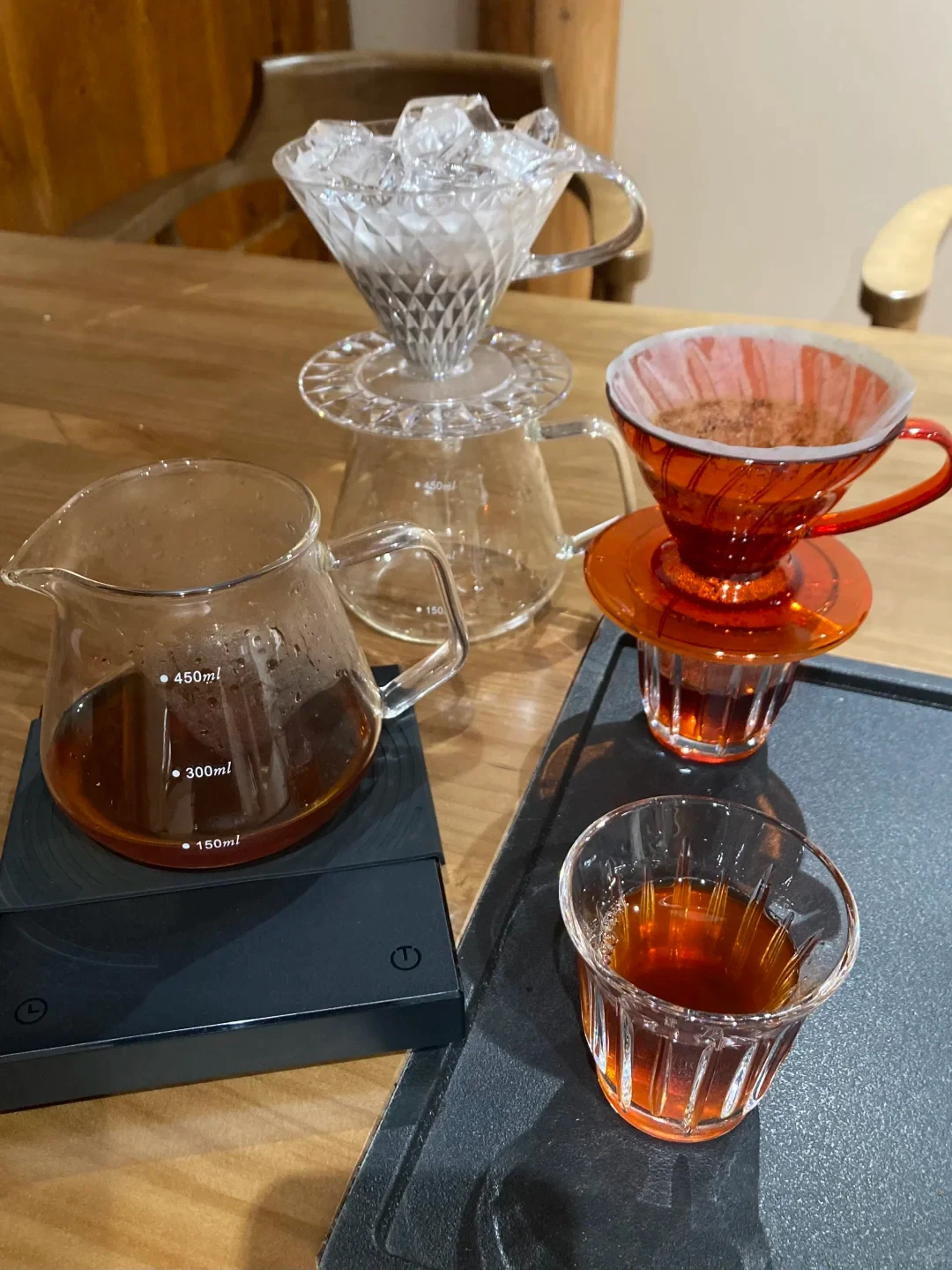 A coffee-brewing setup on a wooden table: a glass carafe with coffee and 150, 300, and 450 ml markings sits on a black digital scale beside a clear dripper holding ice, an amber dripper with coffee grounds, and a glass of brewed coffee.