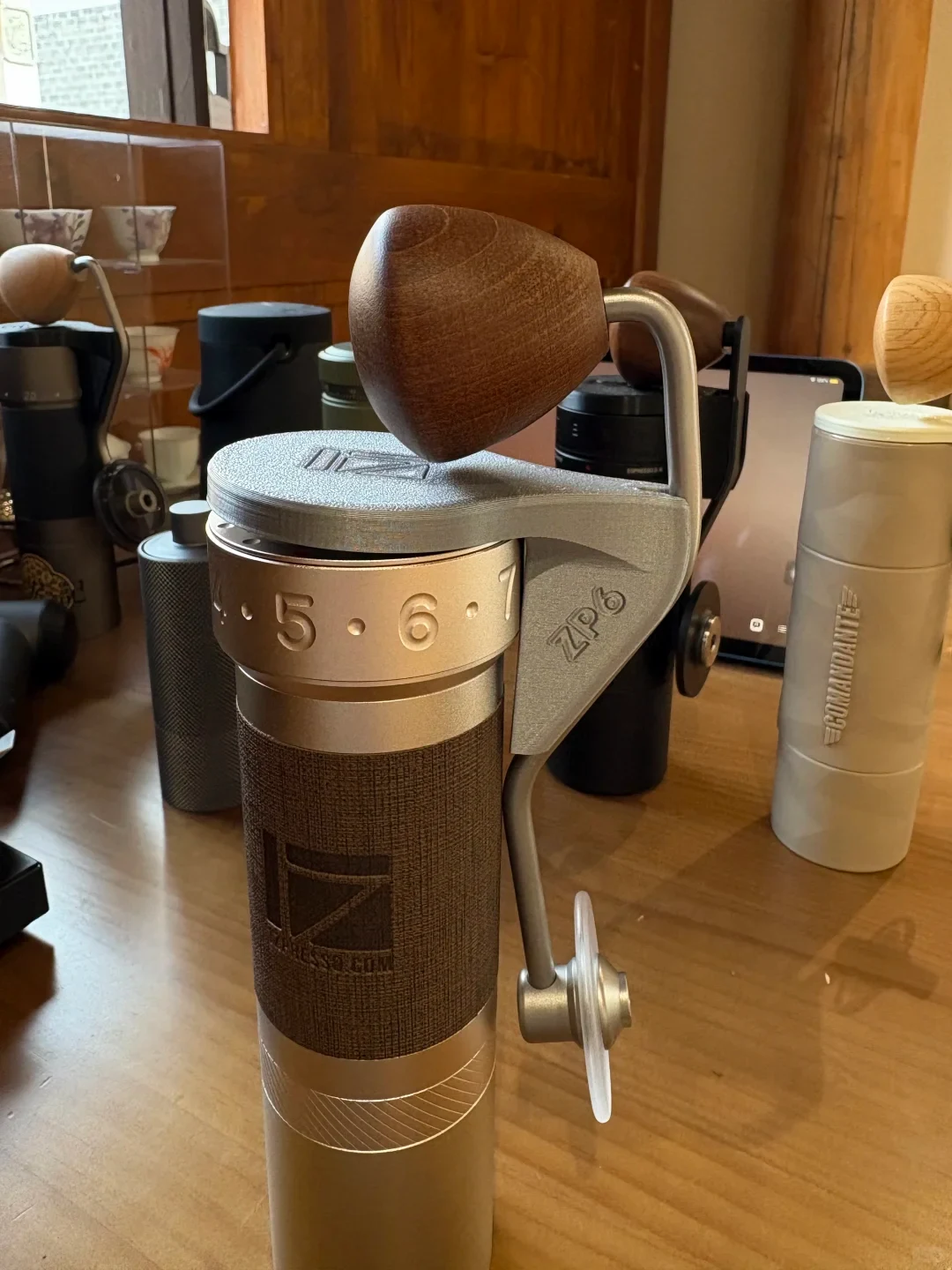 A close-up of a manual coffee grinder on a wooden counter, with a brown wooden crank knob, metal arm, numbered adjustment ring, and textured dark-brown body. Other grinders and containers sit behind it.