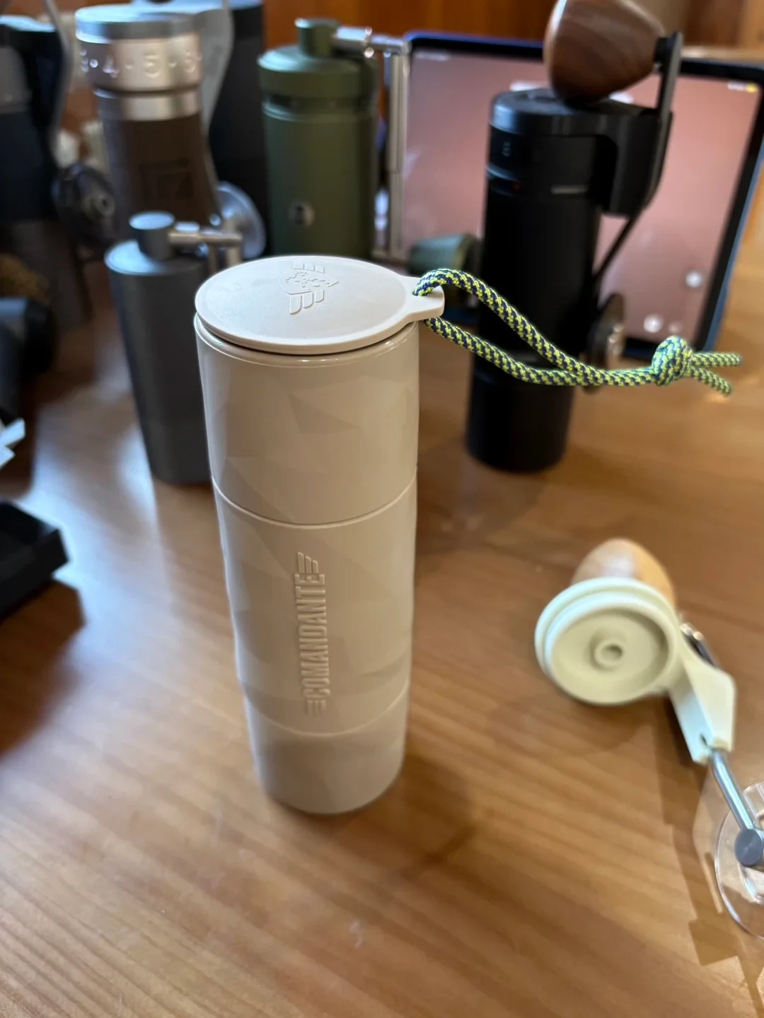 A white COMANDANTE-branded manual coffee grinder has a flat carry lid and a blue-and-yellow cord. Its removed crank and lid lie beside it on a wooden table, with other grinders in the background.