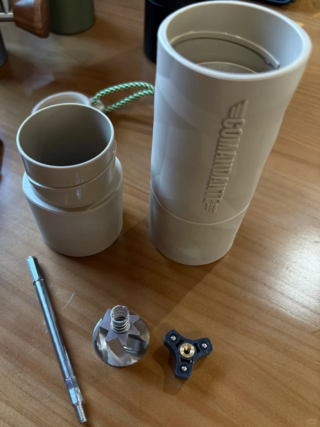 A white cylindrical handheld grinder or mill is shown disassembled on a wooden table, with its open main body and detached cup-like section beside metal grinding parts, a shaft, and a black three-pronged component.