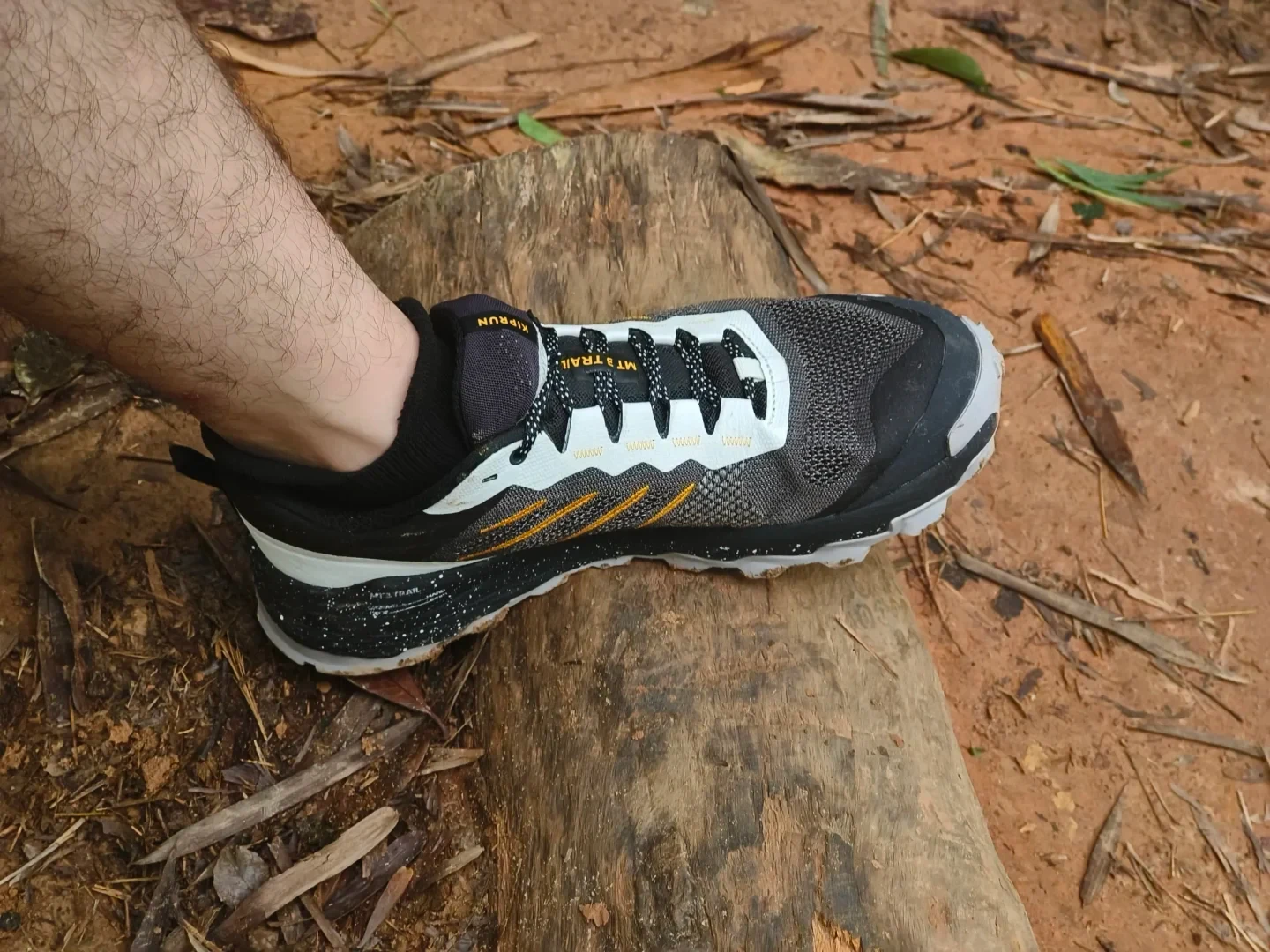 A person's foot wearing a black, grey, and white Kiprun MT3 Trail running shoe resting sideways on a weathered wooden log along a dirt path.