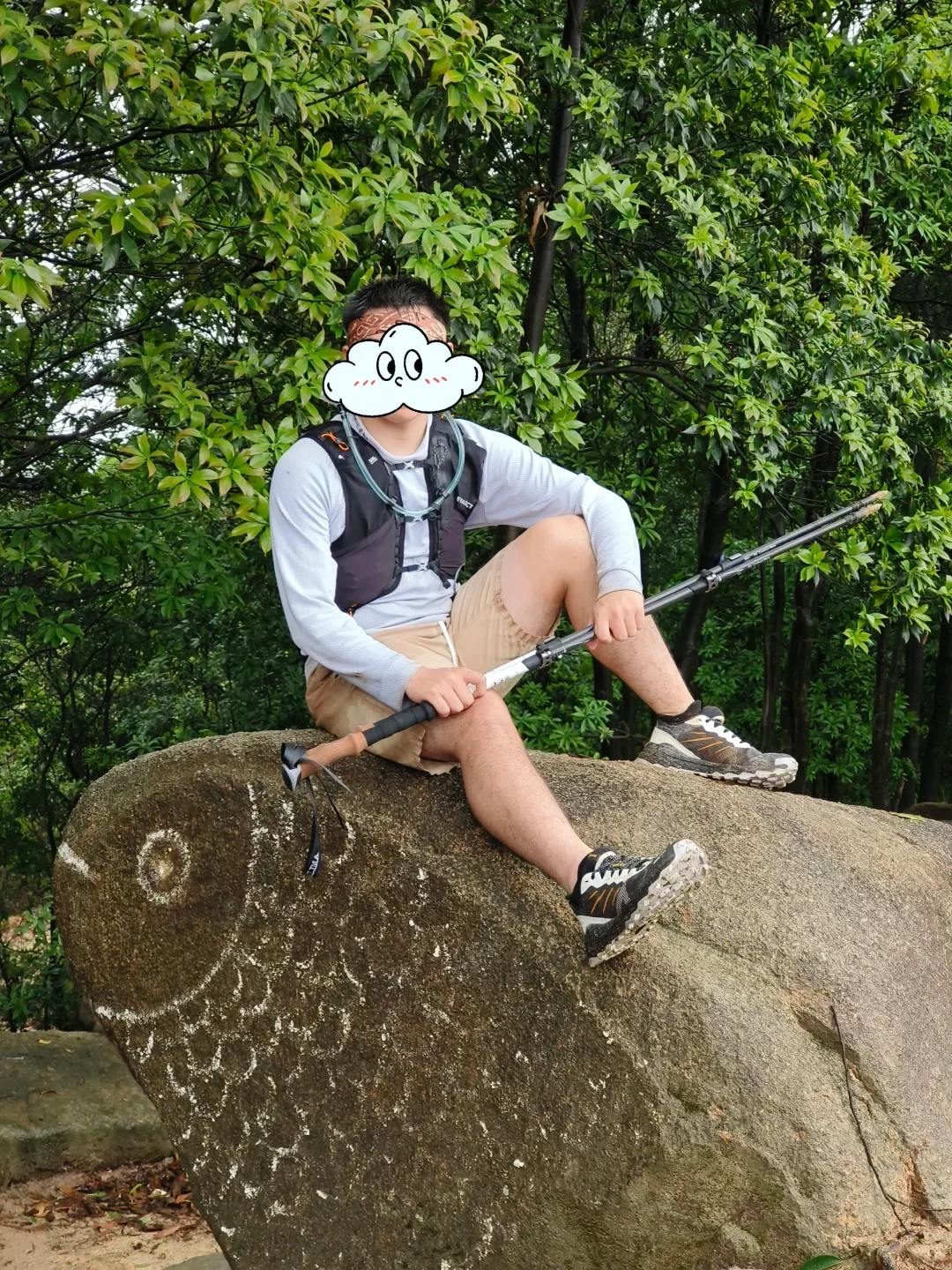 A hiker wearing a light athletic shirt, running vest, and shorts sits on a large boulder etched with a line drawing of a fish. Holding trekking poles across his lap, he poses in front of lush green trees, with his face covered by a cartoon cloud sticker.