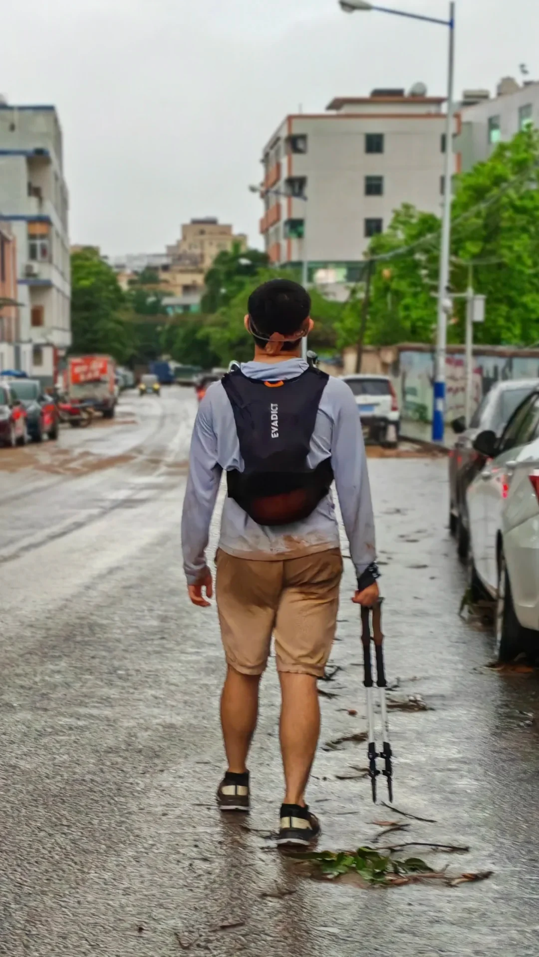 A rear view of a man walking down a wet street lined with buildings and parked cars, wearing a trail running hydration pack and carrying trekking poles, with mud stains on his shirt and shorts.