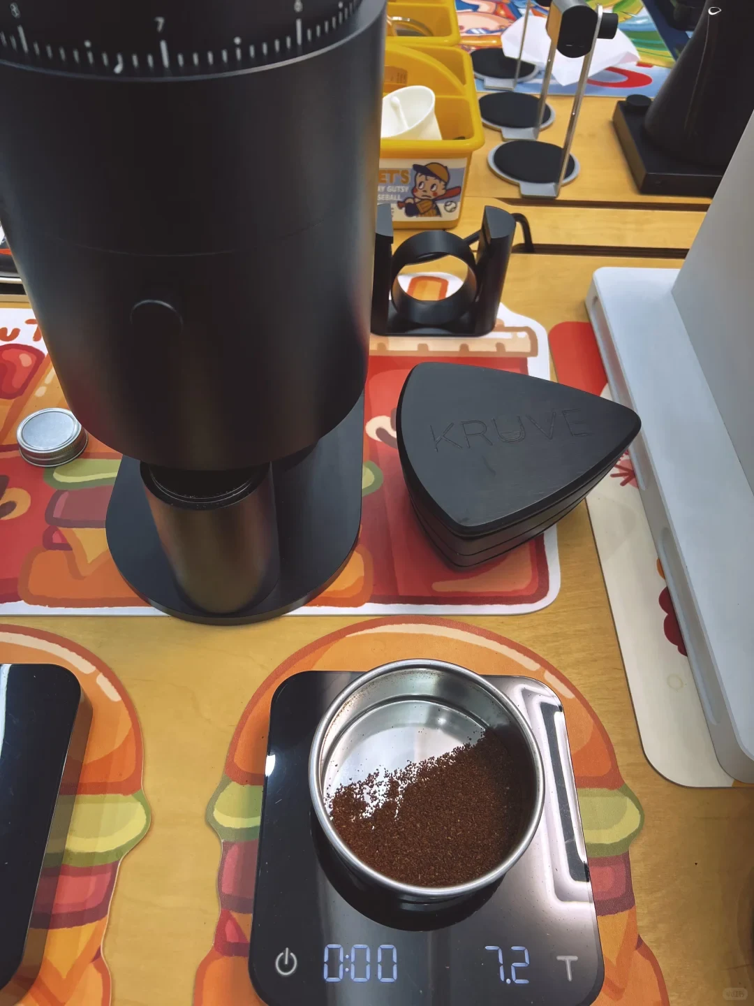 A metal sieve containing coffee grounds rests on a digital scale in front of a black grinder. A triangular KRÜVE sieve lid lies beside the grinder.