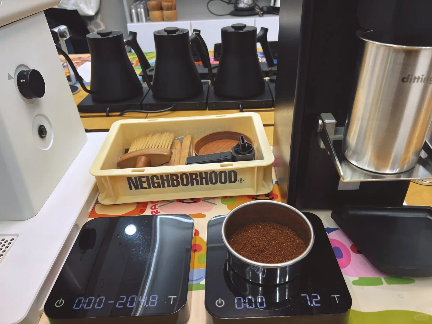 Coffee grounds in a metal sieve sit on a digital scale beside a white grinder. Kettles, brushes and a tall Ditting grounds container are arranged behind it.