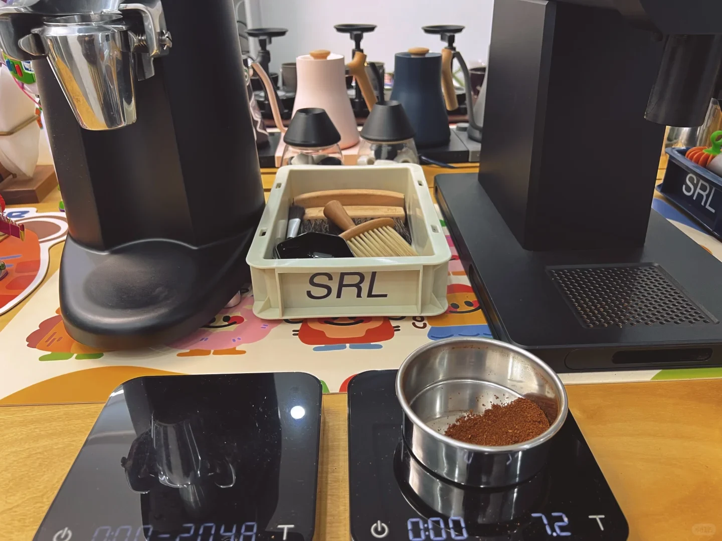 Coffee grounds in a metal sieve rest on a digital scale in front of a black cylindrical grinder. A second black grinder and a basket of brushes stand behind it.