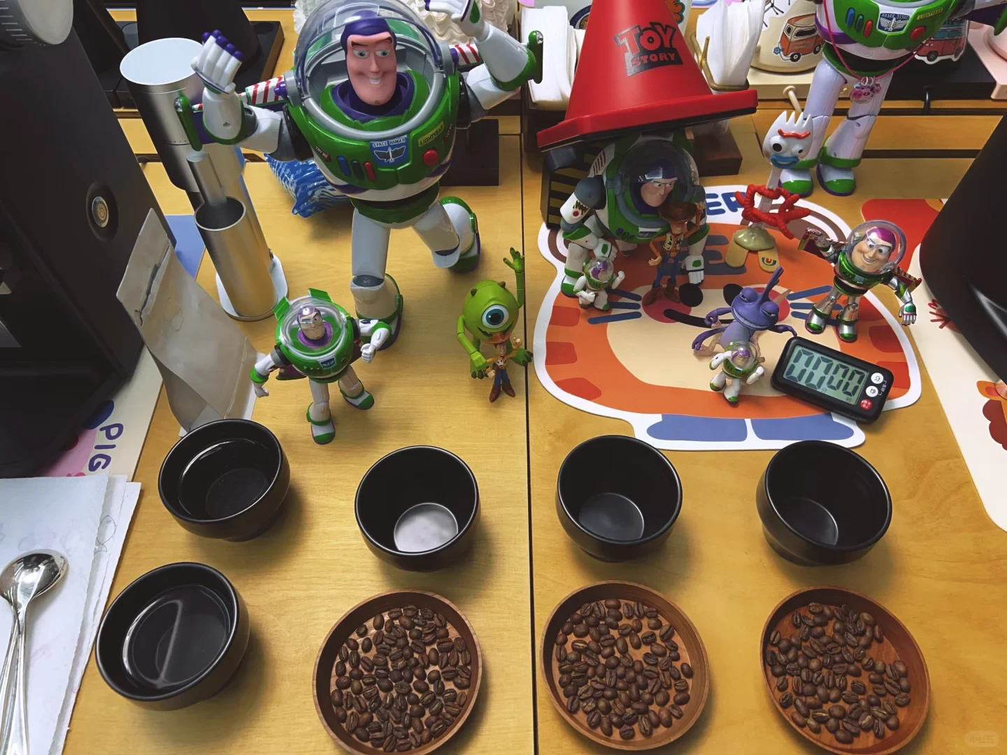 Coffee beans are divided among black cupping bowls on a counter, with a timer and decorative figures behind them.