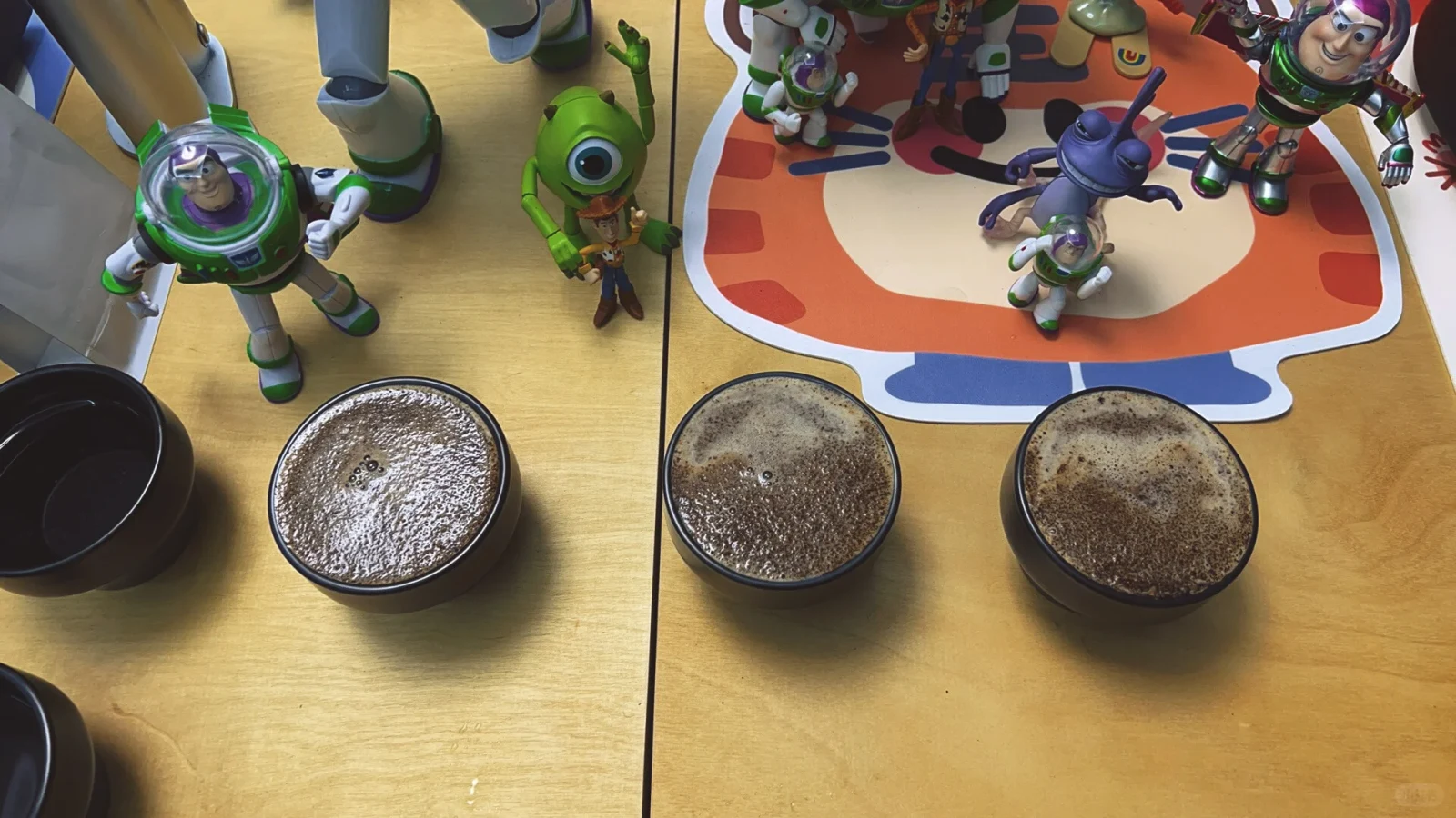 Three cupping bowls hold coffee with patches of foam on the surface; decorative figures stand behind the bowls.