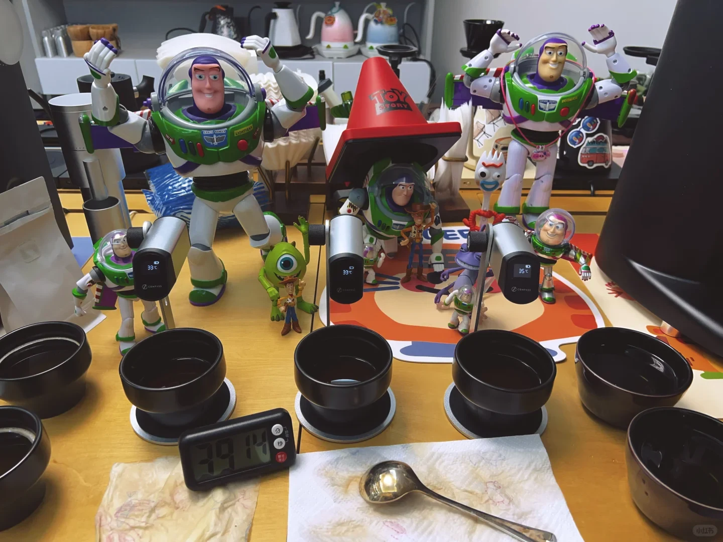 Black cupping bowls, probe thermometers, a spoon, a timer and a small bag of beans are arranged on the counter, with decorative figures behind them.