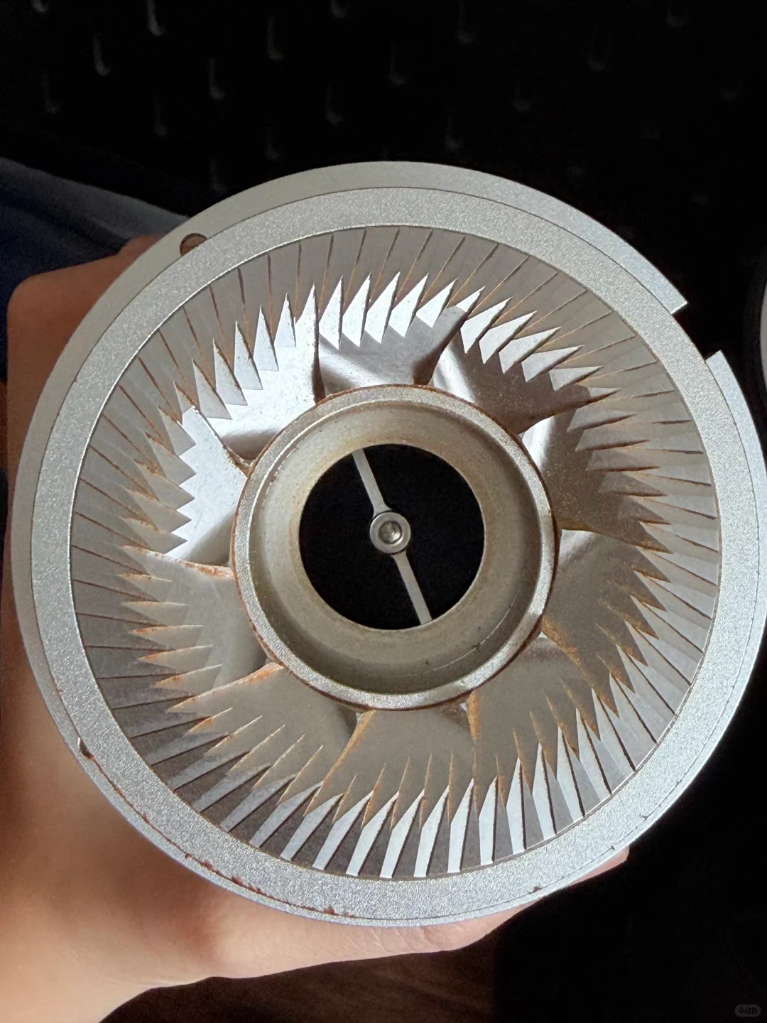 A hand holds the detached upper flat coffee-grinder burr, with brown coffee fines visible in the grooves and around its inner edge.