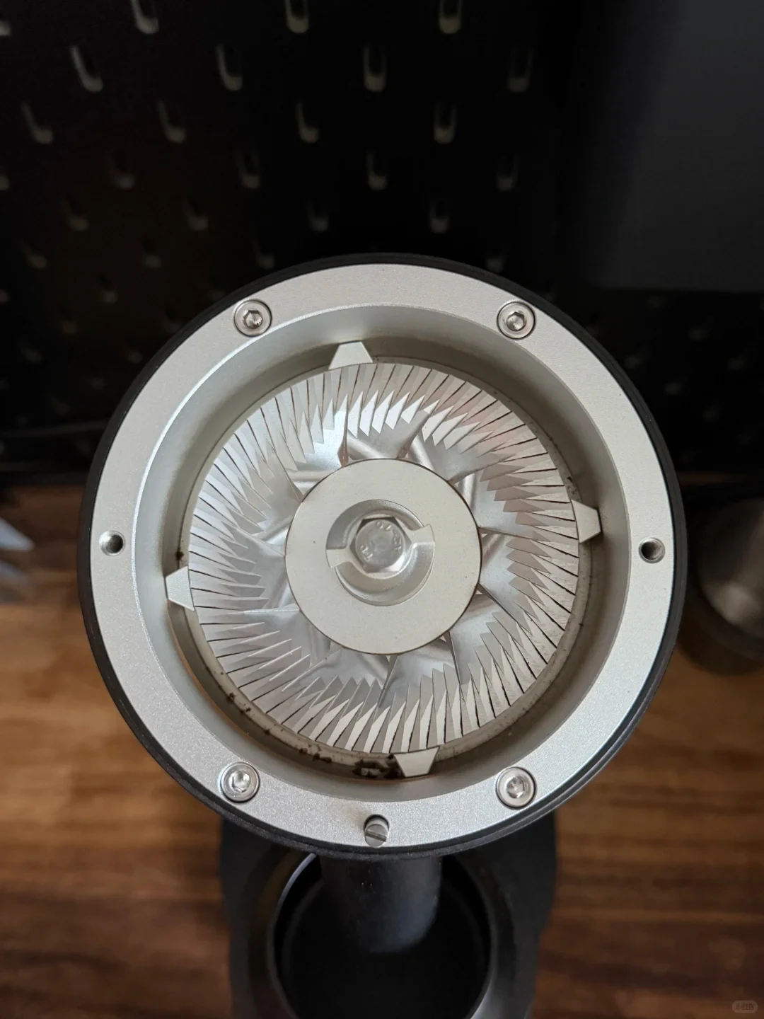 Close-up of a circular metal grinder burr assembly with many radial cutting teeth and a central fastener, mounted on a black appliance above a wooden surface.