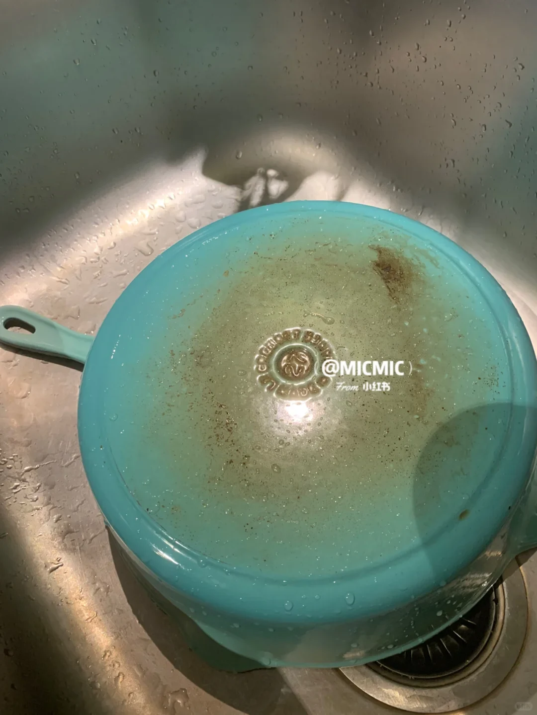 The underside of a wet turquoise enameled pan resting upside down in a stainless steel sink, showing browned burn stains across its base.