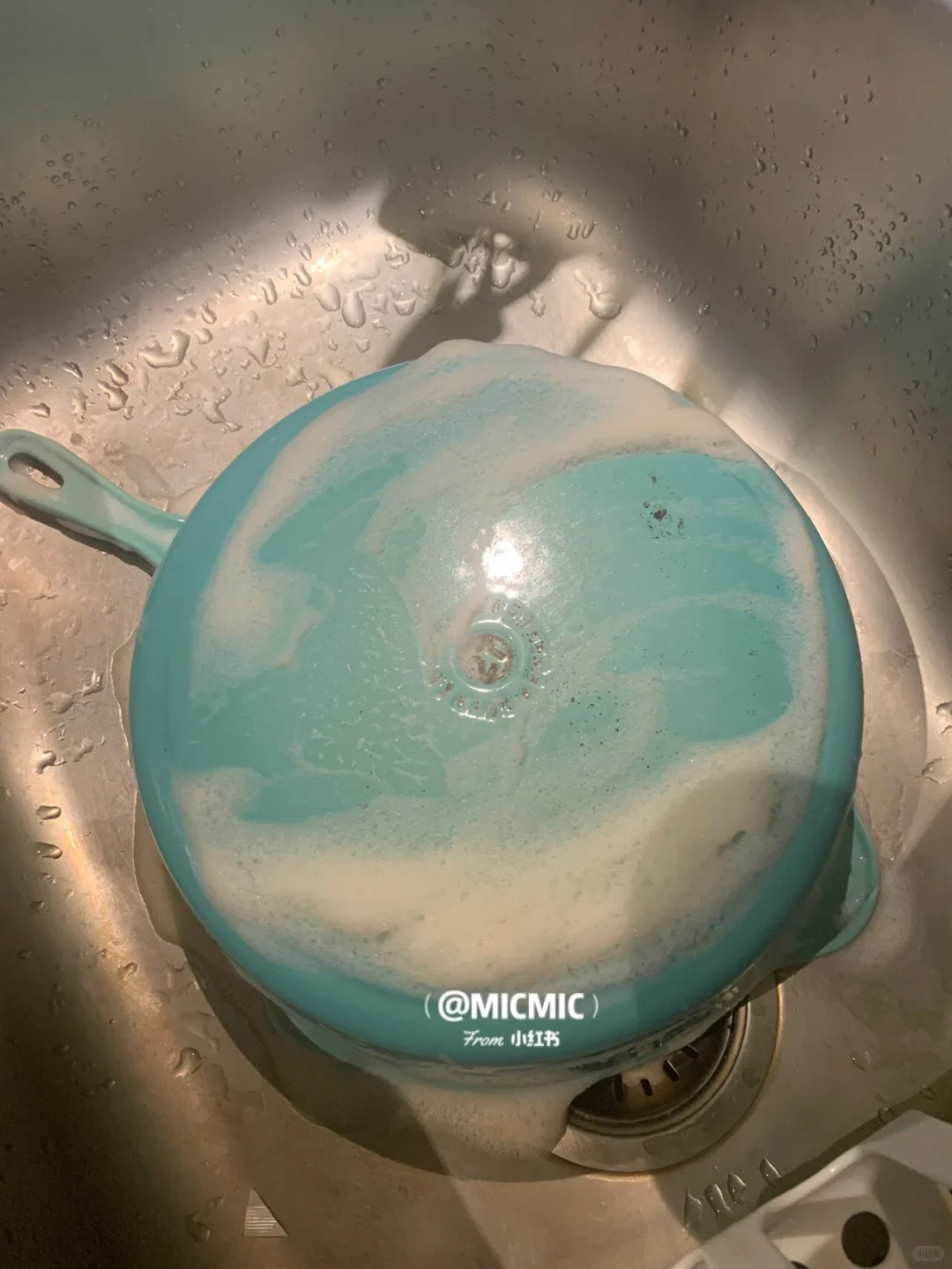 The turquoise Le Creuset pan turned upside down in a stainless steel sink, with white cleaner spread across its exterior base.