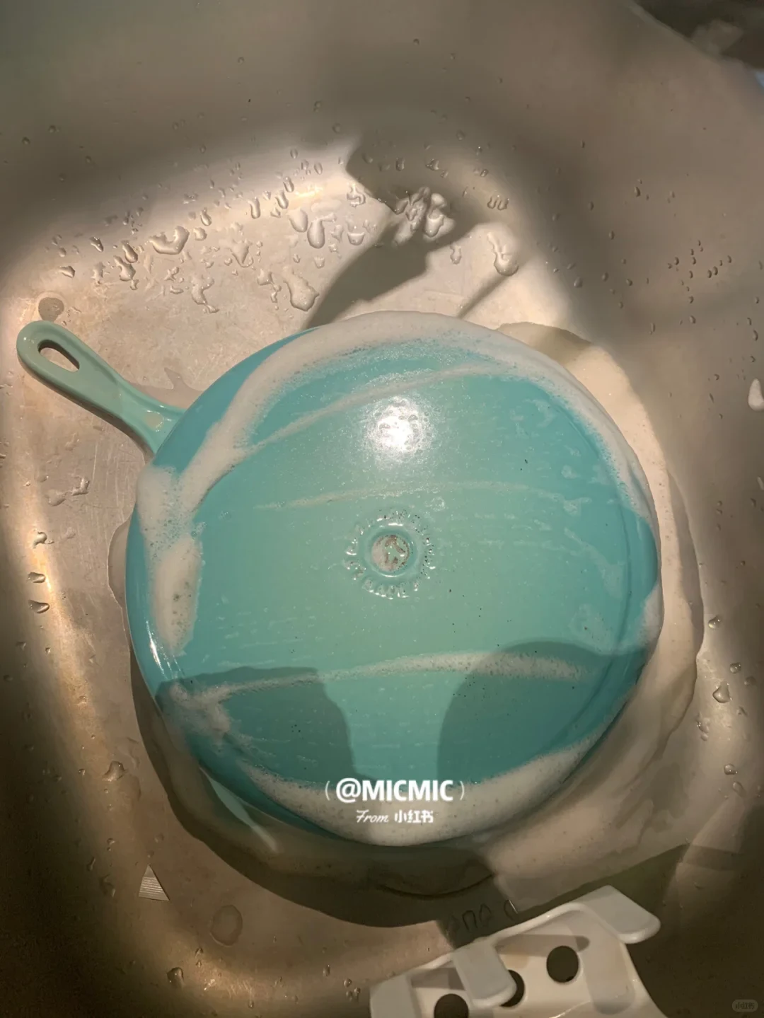 A light blue enameled frying pan placed upside down in a stainless steel sink, covered in white cleaning foam.