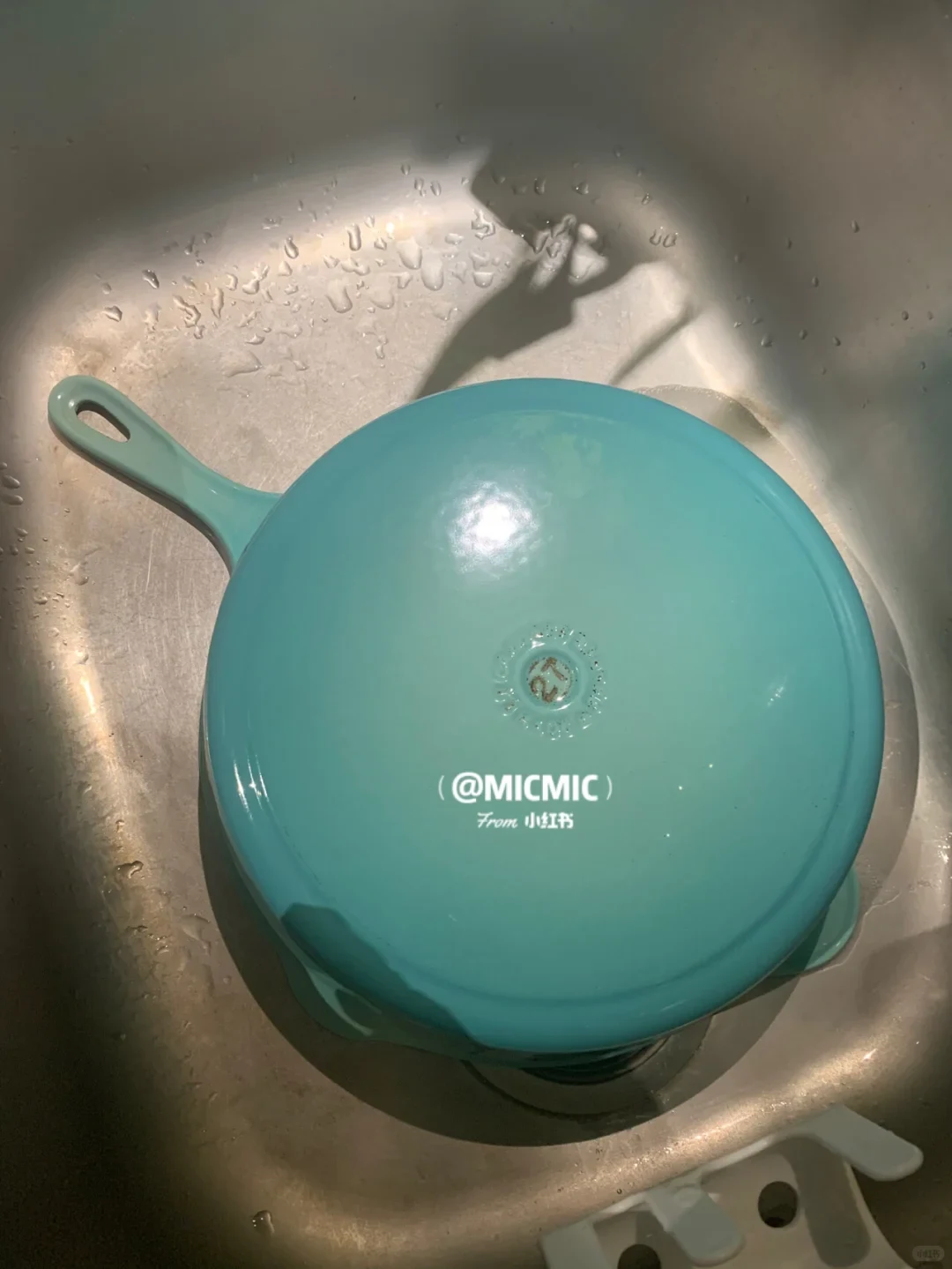 An overturned turquoise enameled cast iron skillet resting in a wet stainless steel sink.