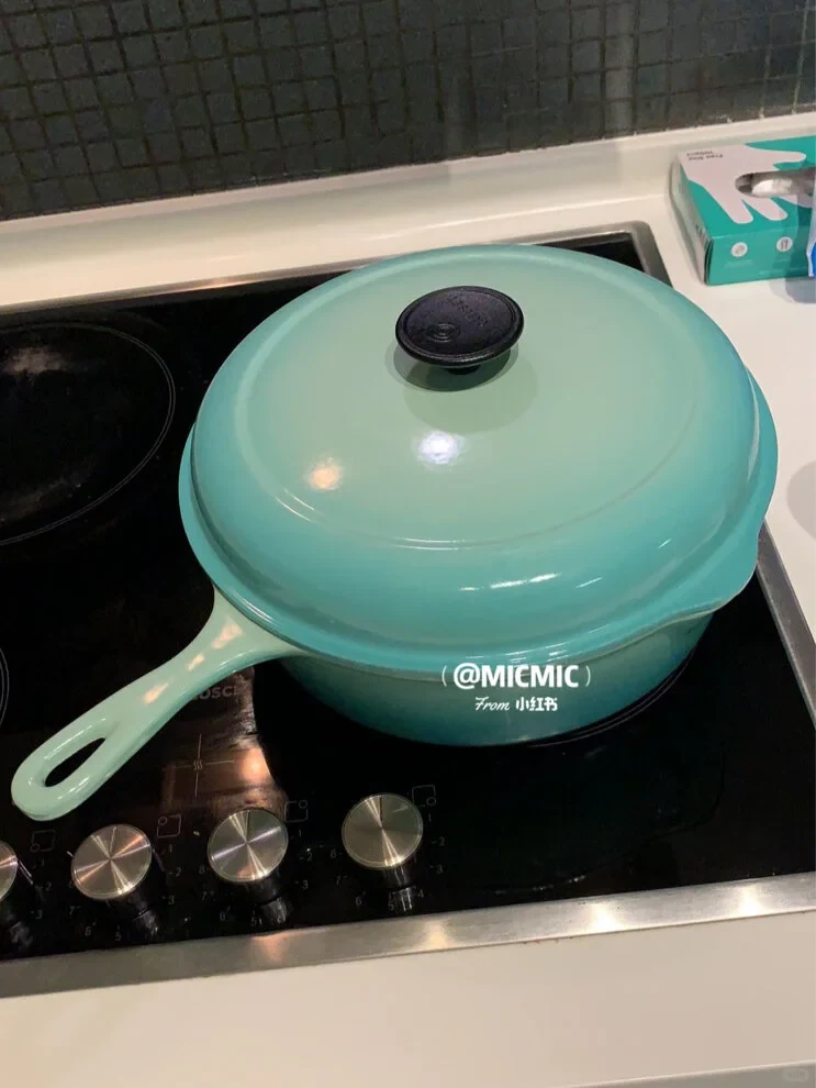 A turquoise Le Creuset deep pan with a long handle and domed lid on a black glass cooktop.