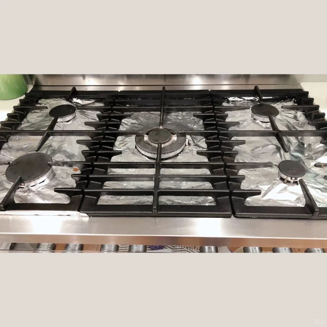 A stainless-steel gas cooktop with five burners and black cast-iron grates; the surface is covered in crinkled foil.