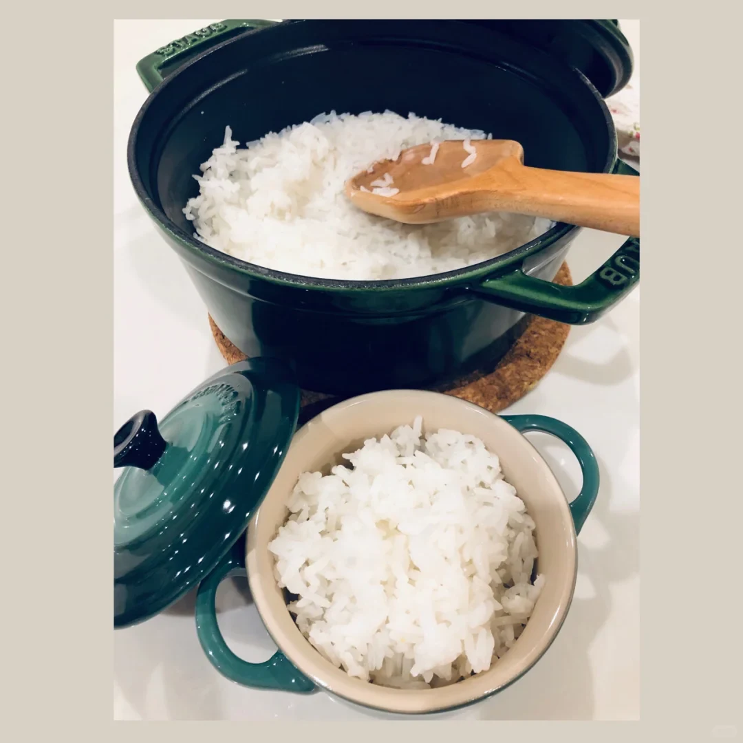 A dark green enameled pot of cooked white rice sits open on a cork trivet, with a wooden rice paddle resting inside; a matching bowl filled with rice and the pot lid are in the foreground.