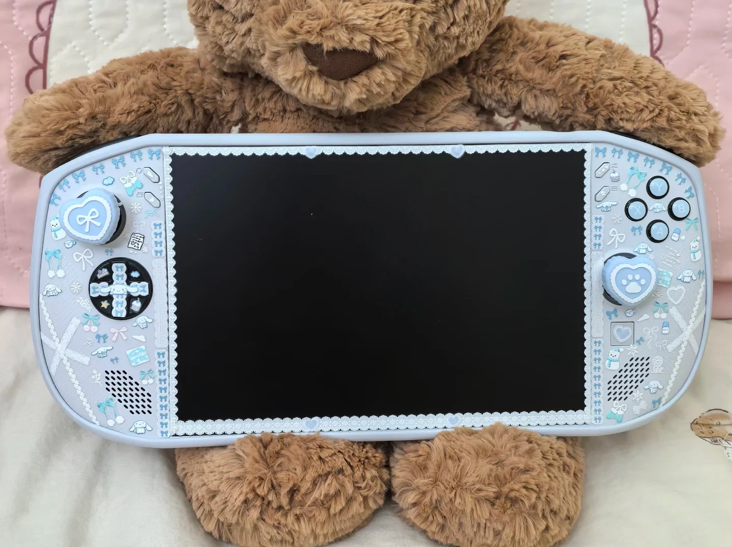 A brown plush teddy bear holding a handheld gaming console decorated with light blue and white lace borders, bows, and cute cartoon stickers on its white body and thumbsticks.
