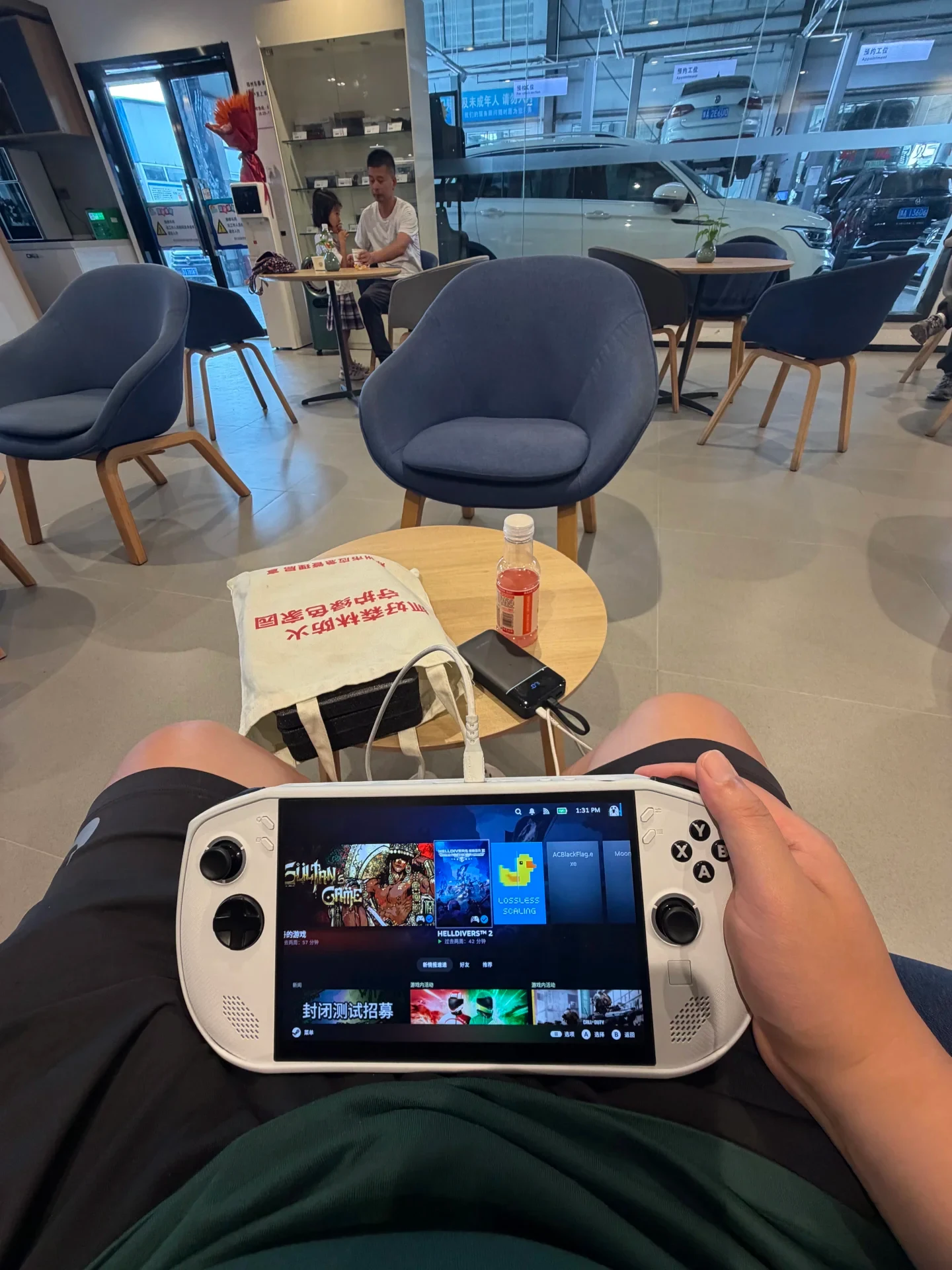 A first-person view of a person holding a white handheld gaming console displaying a game library interface while seated in a waiting lounge. In front of them on a small round table sits a tote bag, a beverage bottle, and a power bank wired to charge the device.