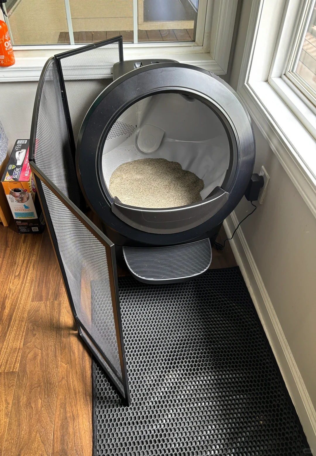 A black Litter-Robot 4 automatic cat litter box set on a black honeycomb litter mat next to a window, flanked on one side by a black mesh fireplace screen.