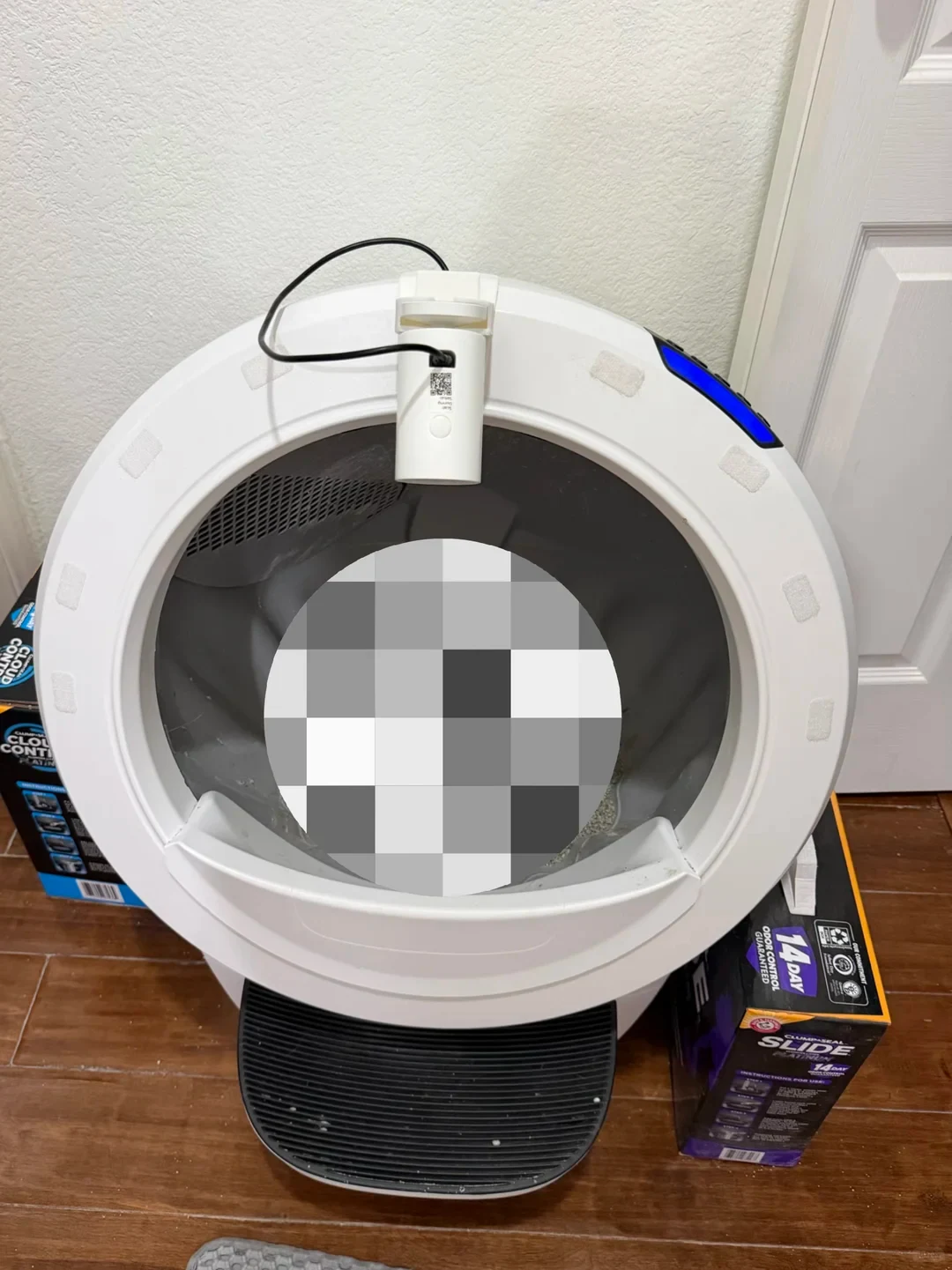 Front view of a white spherical automatic cat litter box placed on a wooden floor, featuring an attached camera or sensor module at the top entrance and a black step grate at the bottom. A large circular pixelated mosaic censors the center of the interior globe, flanked by boxes of cat litter on either side.