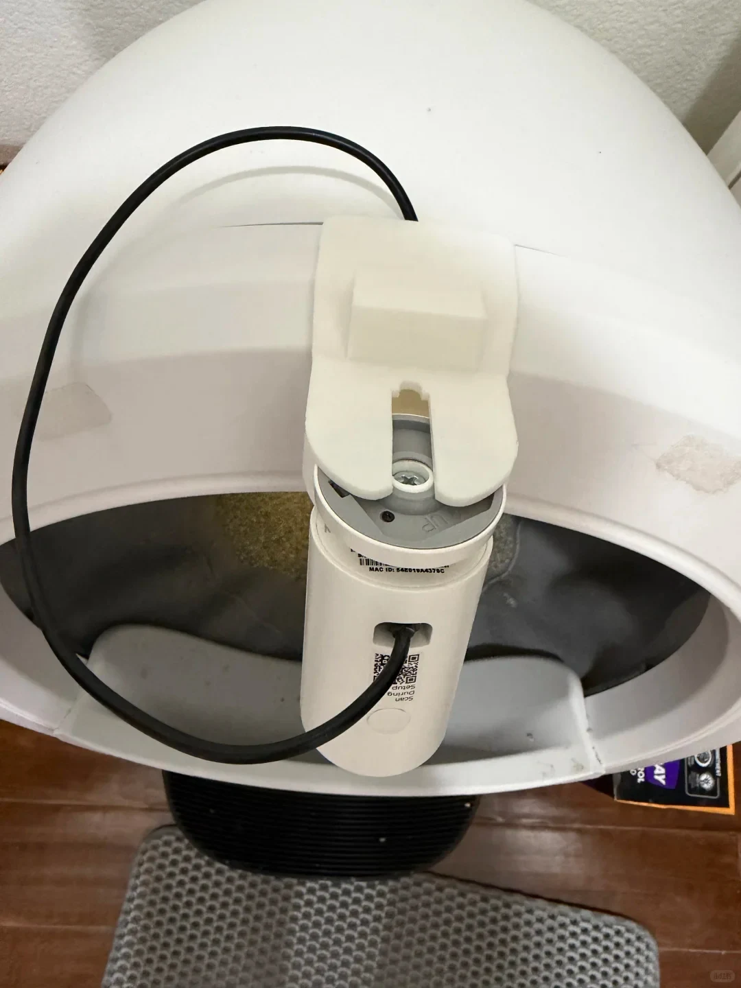 A top-down view of a white automatic cat litter box featuring a cylindrical white accessory with a black cable mounted over the entrance rim with a bracket.