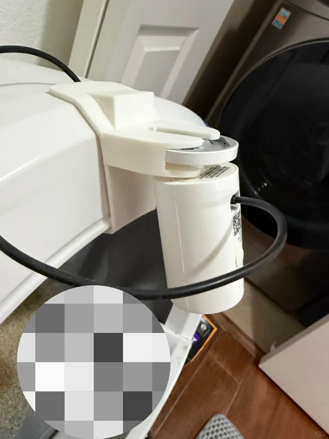A close-up shot of a small white cylindrical camera mounted upside down in a white 3D-printed plastic bracket clamped onto the side of a white household appliance. A black power cable loops around the camera, and a portion in the lower left corner is pixelated. A dark front-load washing machine is visible in the background.