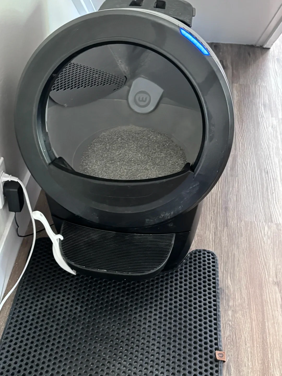 A black Litter-Robot 4 automatic cat litter box plugged into a wall outlet, set up on a wood floor with a black honeycomb litter mat in front of its entry step.