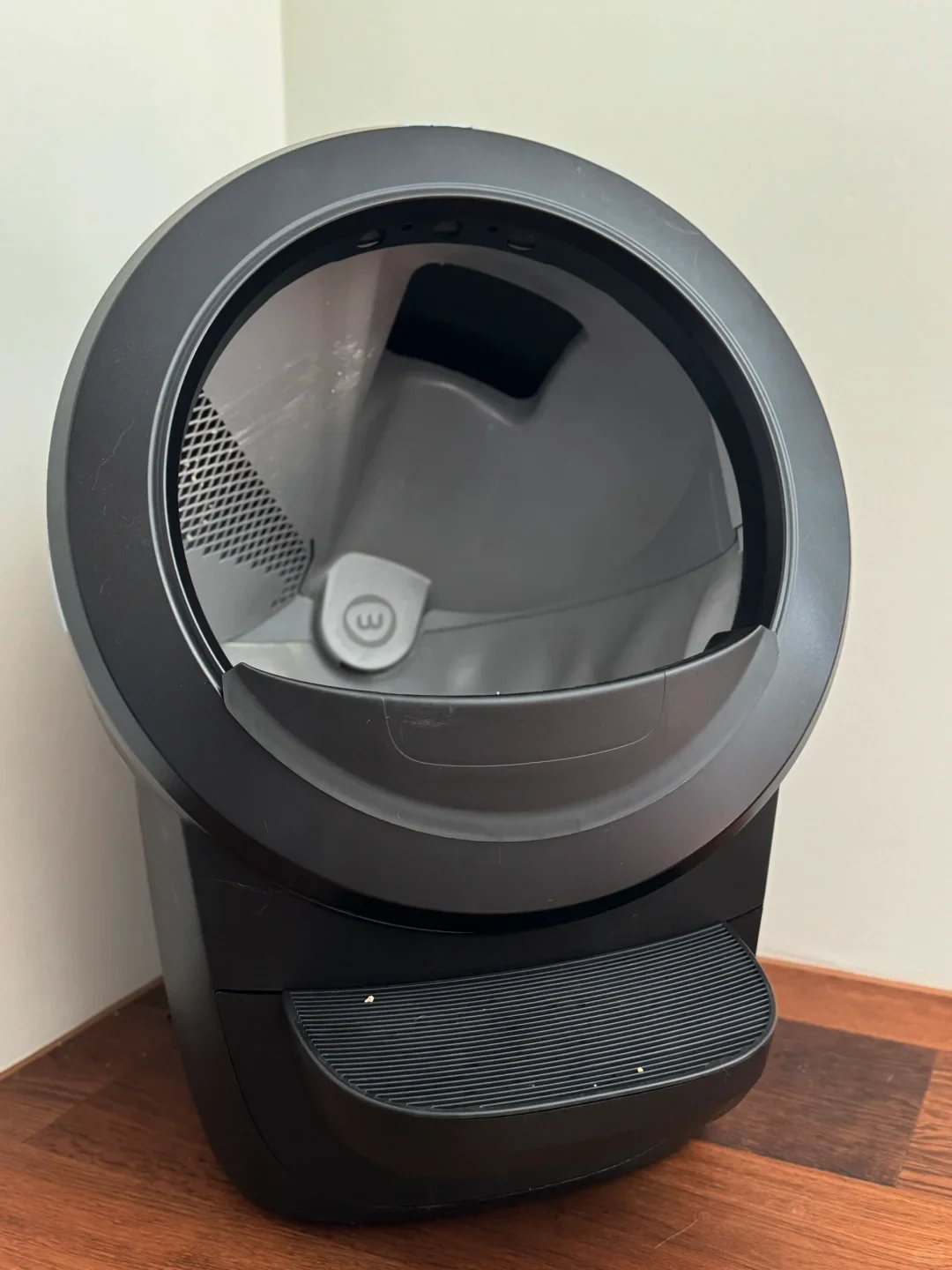 A black automatic self-cleaning cat litter box situated in a corner on a wooden floor, featuring a large circular entrance revealing the gray interior globe and a textured step at the front base.