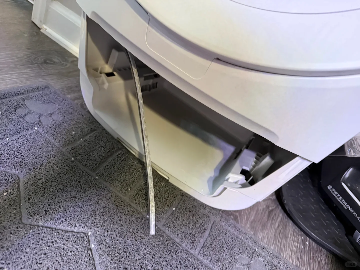 The open waste-drawer compartment of a white automatic cat litter box beside a grey litter mat, with a long narrow detached strip hanging onto the mat.